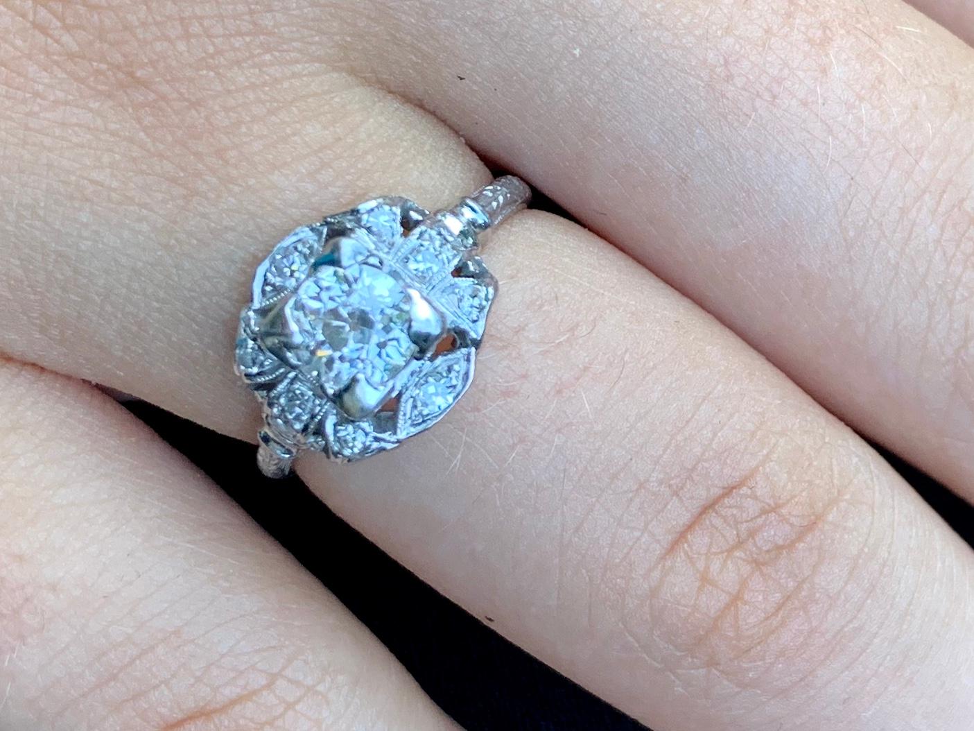Dazzling Art Deco period platinum and diamond engagement ring 
.60 carat central stone surrounded by 10 diamonds for a harmonius, beautiful effect, TCW .95 carat. 
Lovely setting with foliate detail on the sides of the band and unusual detail of two