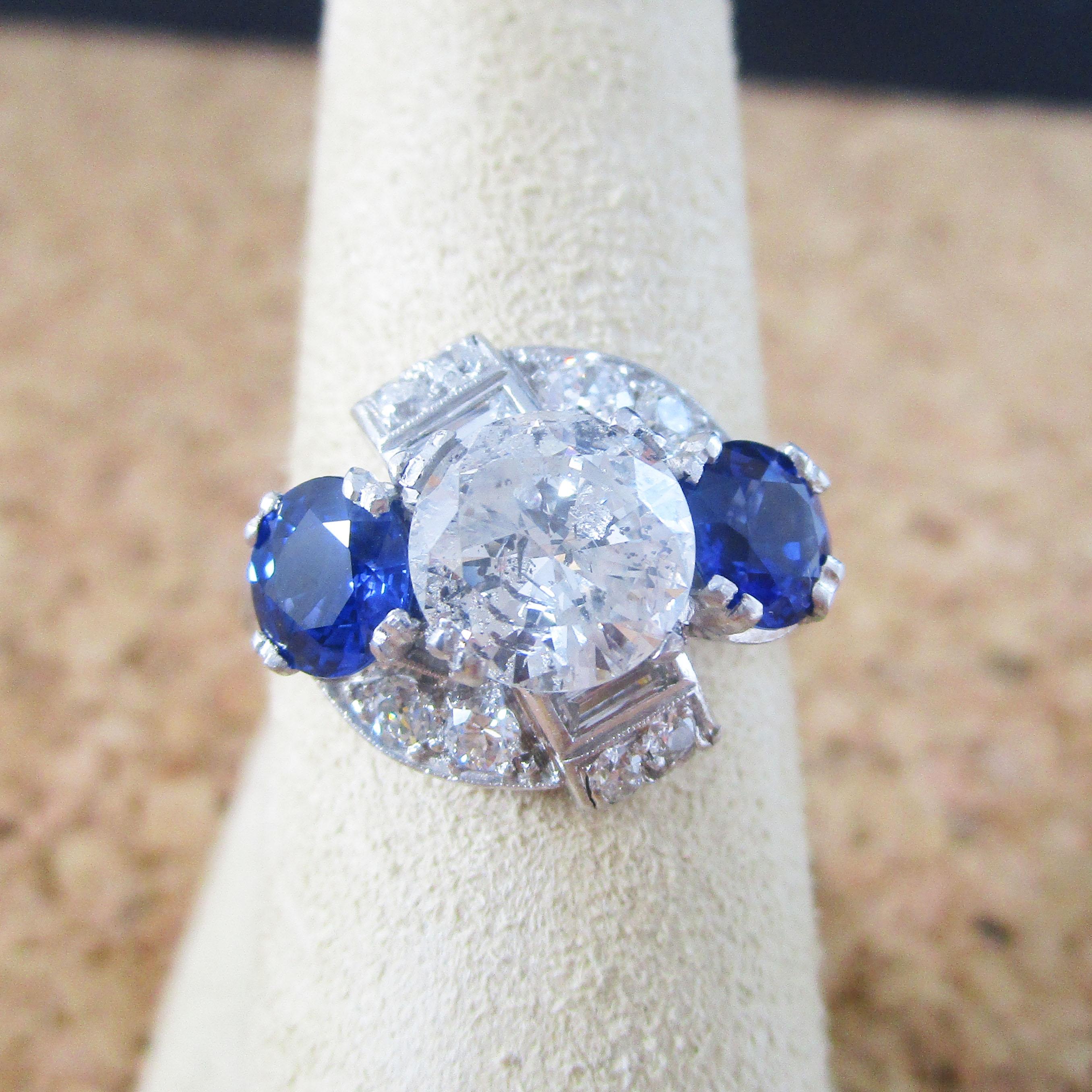 This absolutely breathtaking Art Deco ring is in bright platinum and features a killer 1.75 carat brilliant white diamond center flanked by two gorgeous royal blue sapphires. The ring is incredibly well made and the platinum setting is an all
