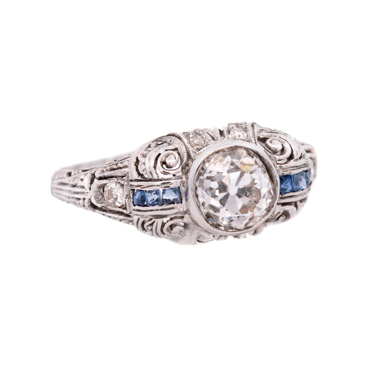 An absolutely beautiful diamond and sapphire engagement ring from the Art Deco (ca1930s) era! Crafted in platinum, this ring features a central, 1.03ct old European cut diamond, which rests within a smooth bezel setting. The diamond displays J color