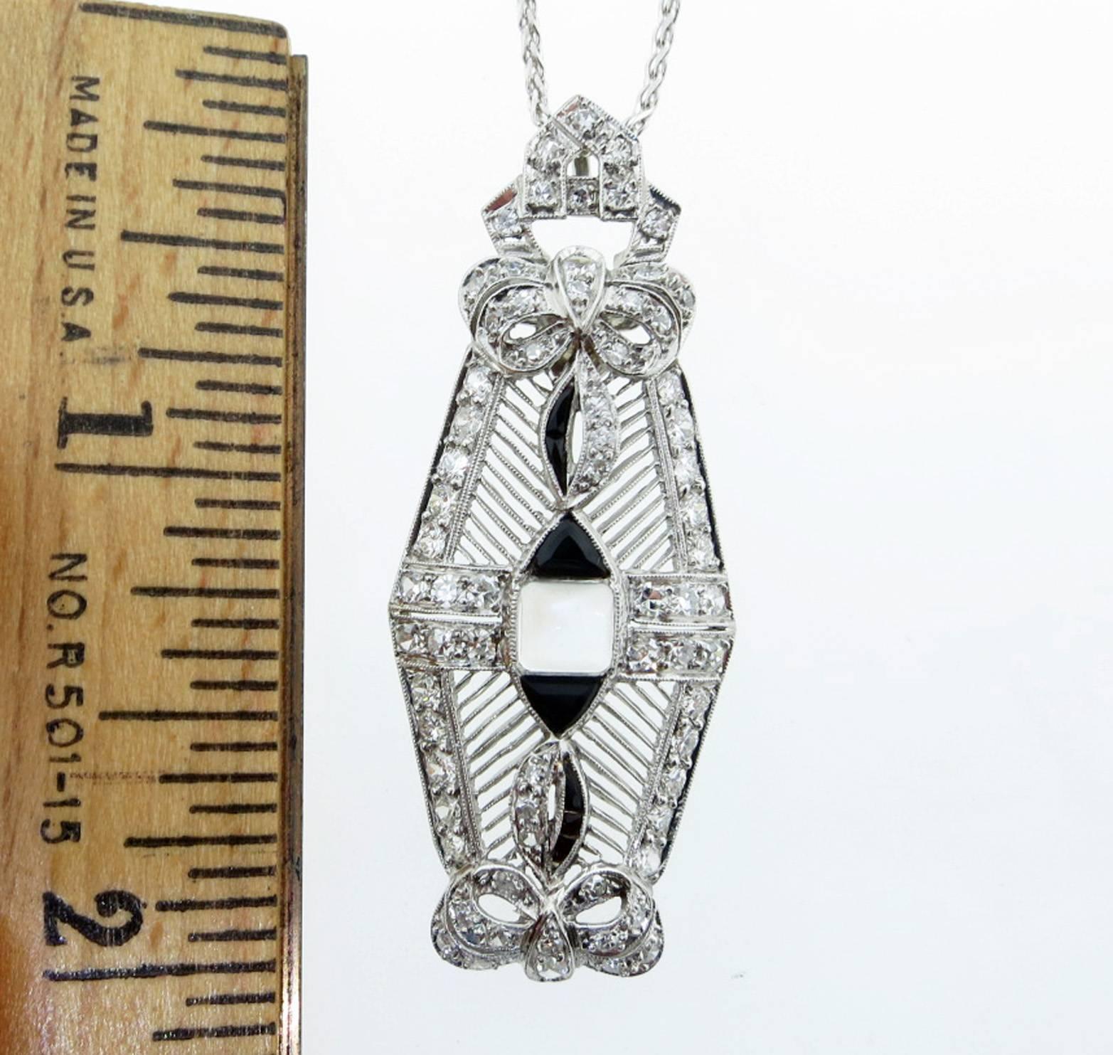 Platinum open work bow motif pendant set with a center pyramid shape moonstone, black onyx accents and 70 round European cut diamonds totaling approx. 1.5cts. The pendant measures approx 2 inches in length  with an articulated pendant top. Circa