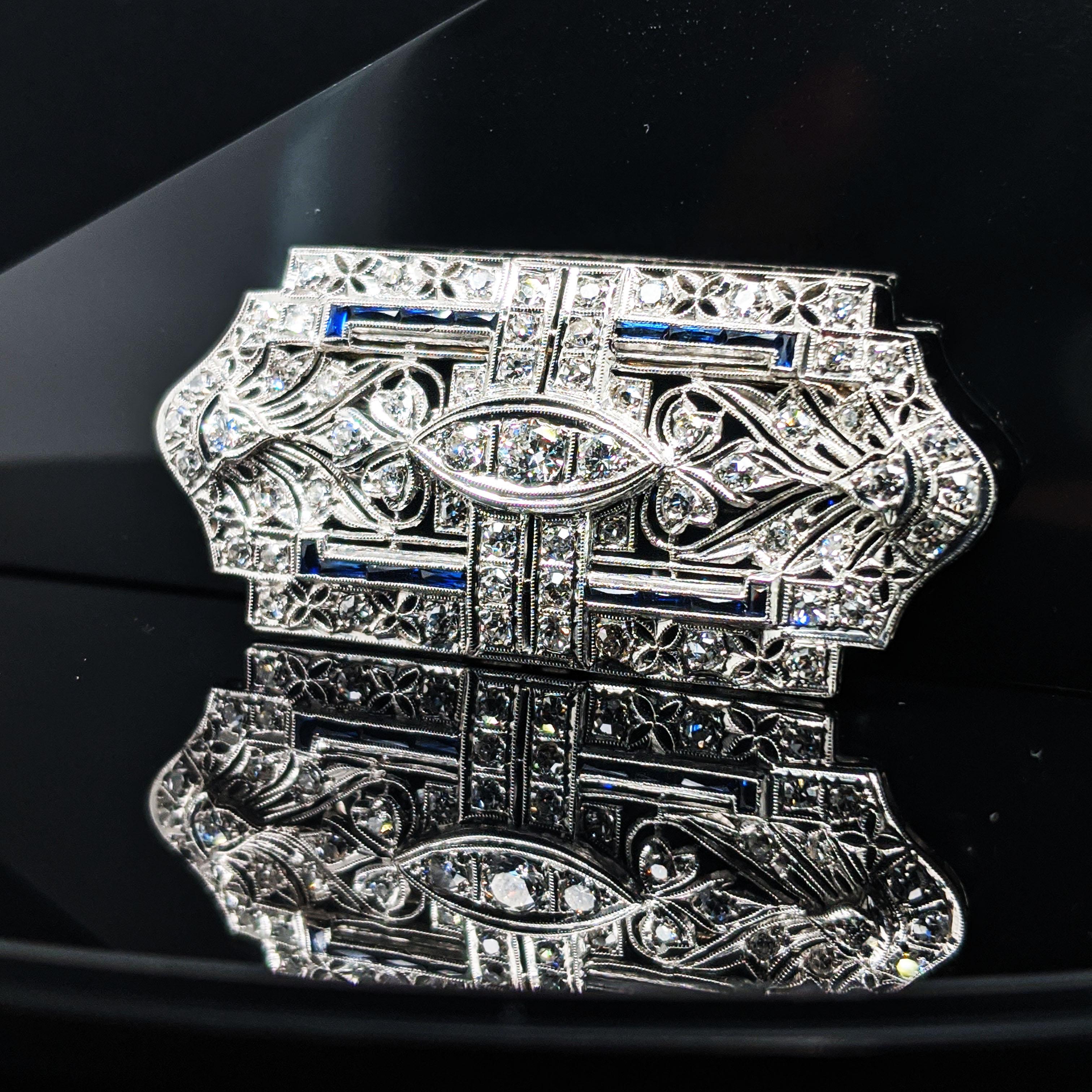 This is a stunning Art Deco pin pendant in platinum with a gorgeous array of bright white diamonds and beautiful blue sapphires. The pin pendant has an intricate pierced design that is accented with fine milgrain detailing. The negative space in the