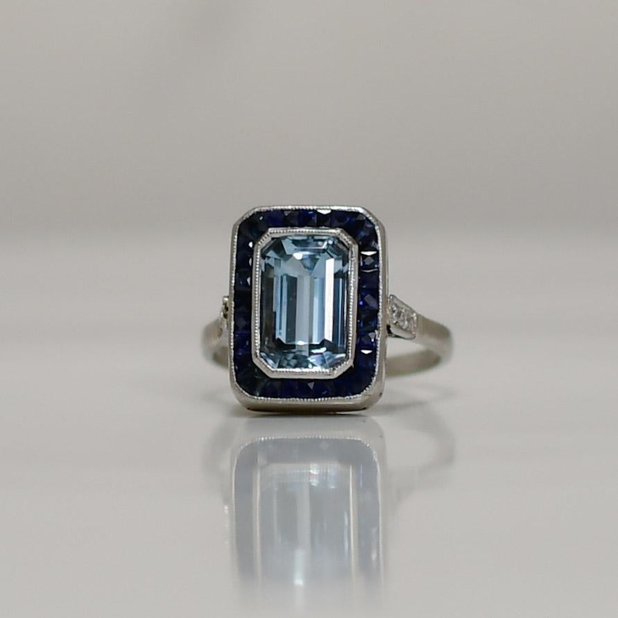 Indulge in the epitome of luxury and elegance with this Platinum Art Deco Emerald Cut Aqua Ring. Its centerpiece showcases a mesmerizing emerald-cut aquamarine, exuding a serene blue hue reminiscent of tranquil waters. The striking sapphire halo and