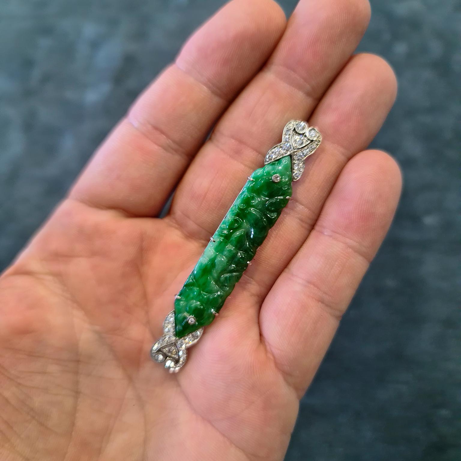 A fine and impressive Art Deco period brooch fashioned in a pierced bar of platinum, mounted with brilliant-cut diamonds at each end, with a carved green jadeite centre panel depicting fruits. Total diamond carat weight of approximately 1.25 carats.