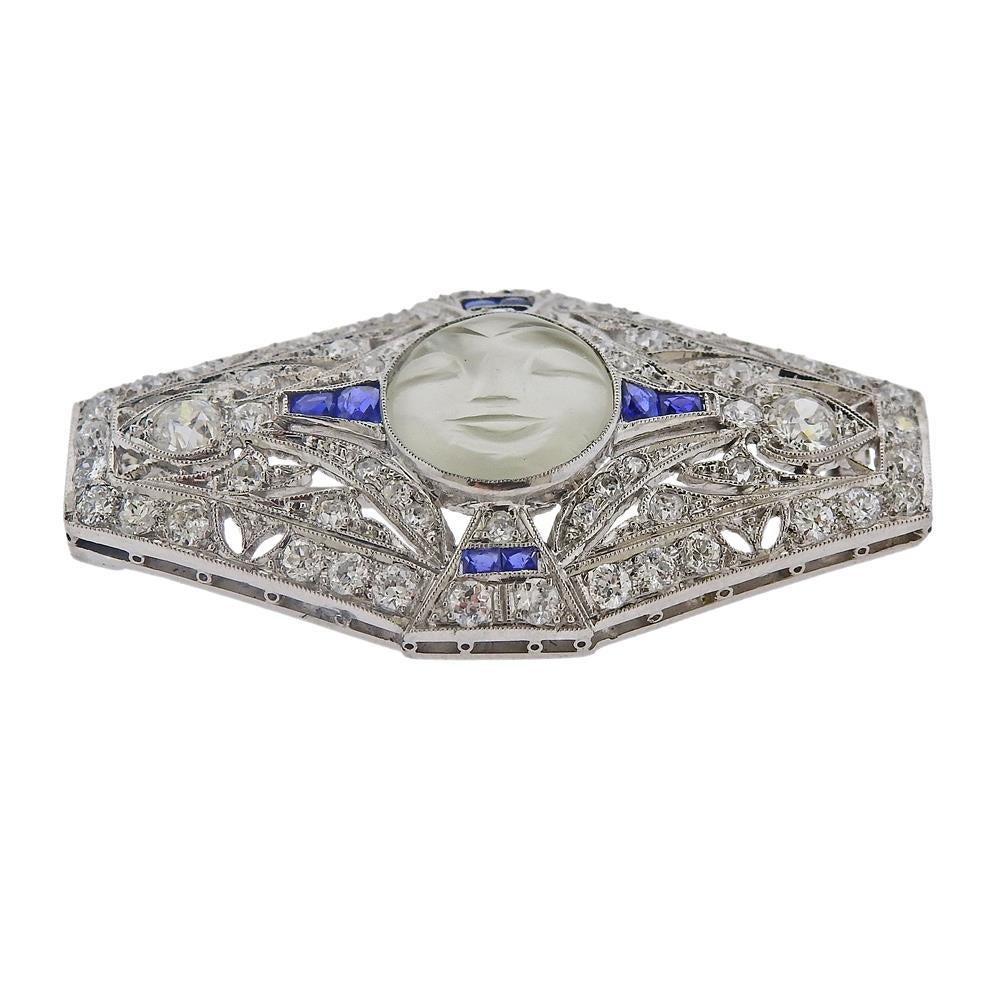 Platinum brooch, set with center carved moonstone, depicting a moon face, surrounded with sapphires and approx. 1.25ctw in Old European cut diamonds. Brooch is 42mm x 23mm. Weighs 11.1 grams.