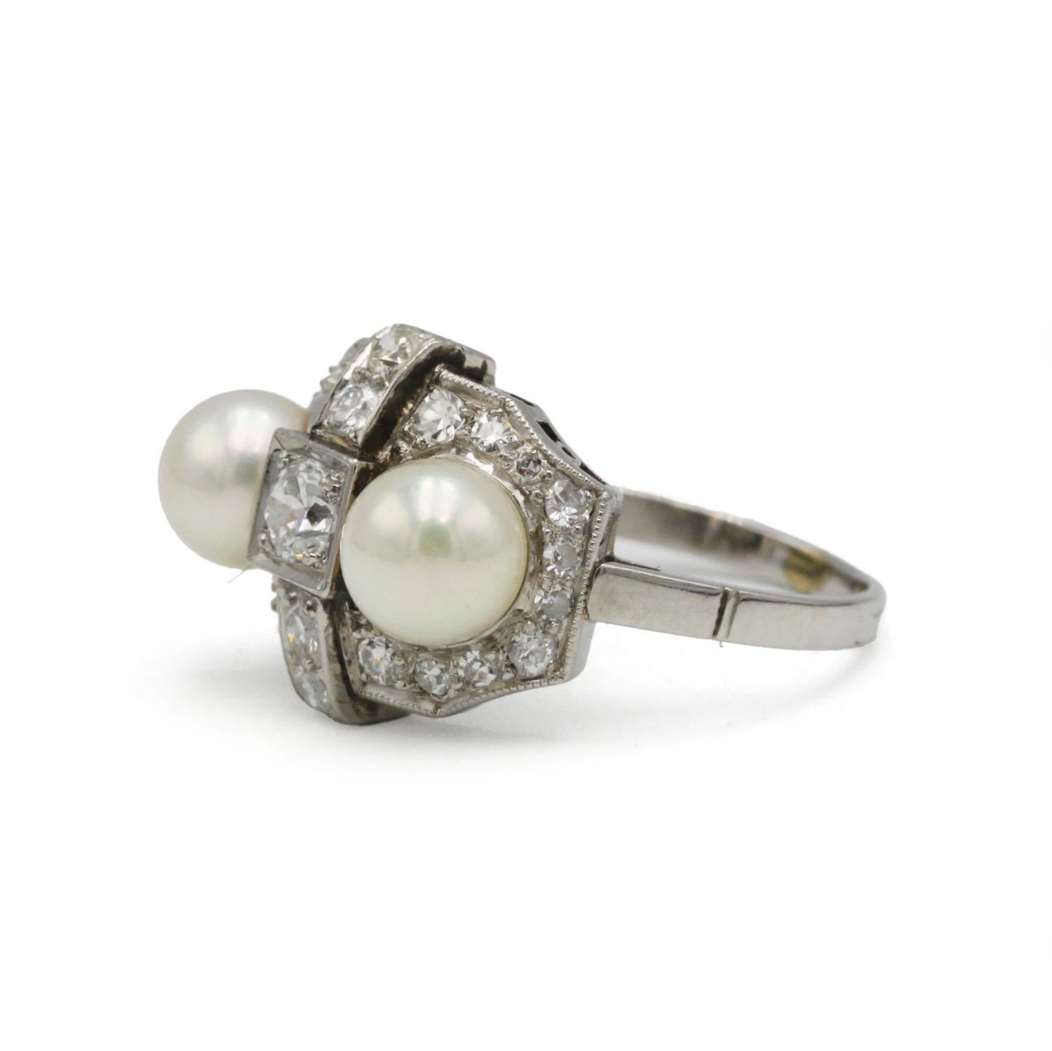 Dieser elegante Platinring im Art-déco-Stil zeigt eine raffinierte und sehr architektonische Komposition, in deren Mittelpunkt ein kreuzförmiges Diamantmotiv steht, das von zwei Perlen mit schönem, gleichmäßigem Glanz eingerahmt wird. Der