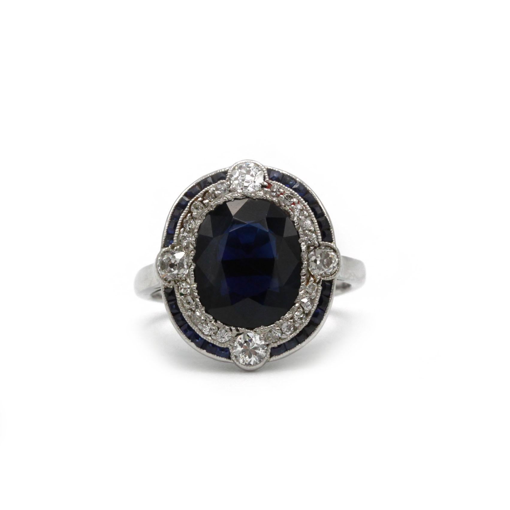 Superbe bague Art-Déco en platine, centrée sur un saphir ovale d'un bleu profond d'environ 3,5 carats. La pierre est entourée d'un délicat halo de saphirs carrés calibrés et d'un second halo intérieur festonné de diamants pavés de taille ancienne,