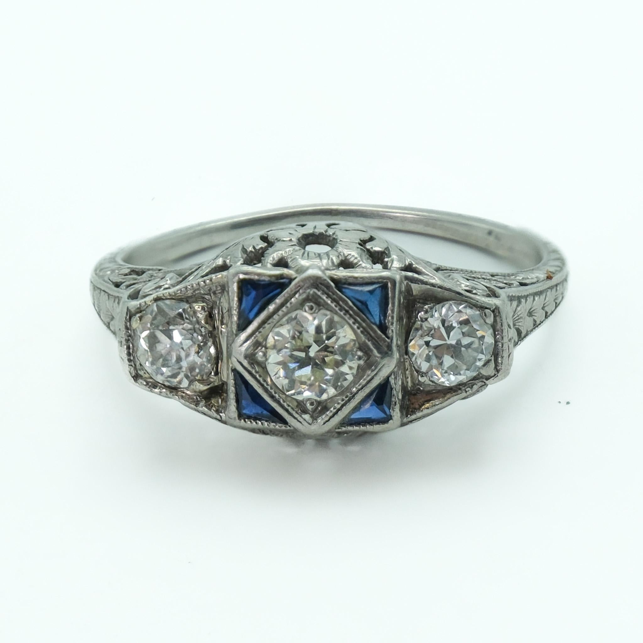 Beautiful handcrafted platinum art deco filligree three stone diamond and synthetic sapphire ring. This historic ring was created circa 1920s and is in fine vintage condition. Carved on the sides with detailed scroll work adding a fine layer of