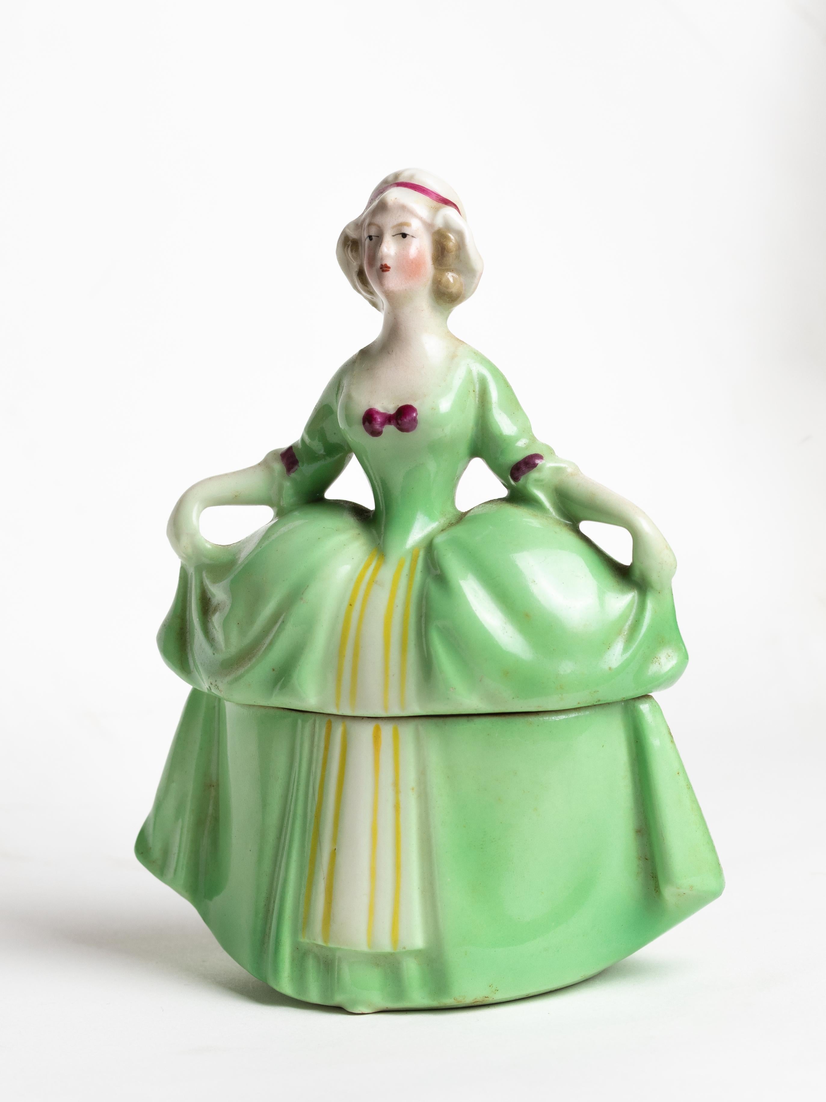 An exquisite Art Deco porcelain jewelry box from 1920, designed as doll portraying Madame Pompadour in a flowing green gown. This charming figure opens to reveal a hidden compartment, originally used to store rings, earrings, or small vanity
