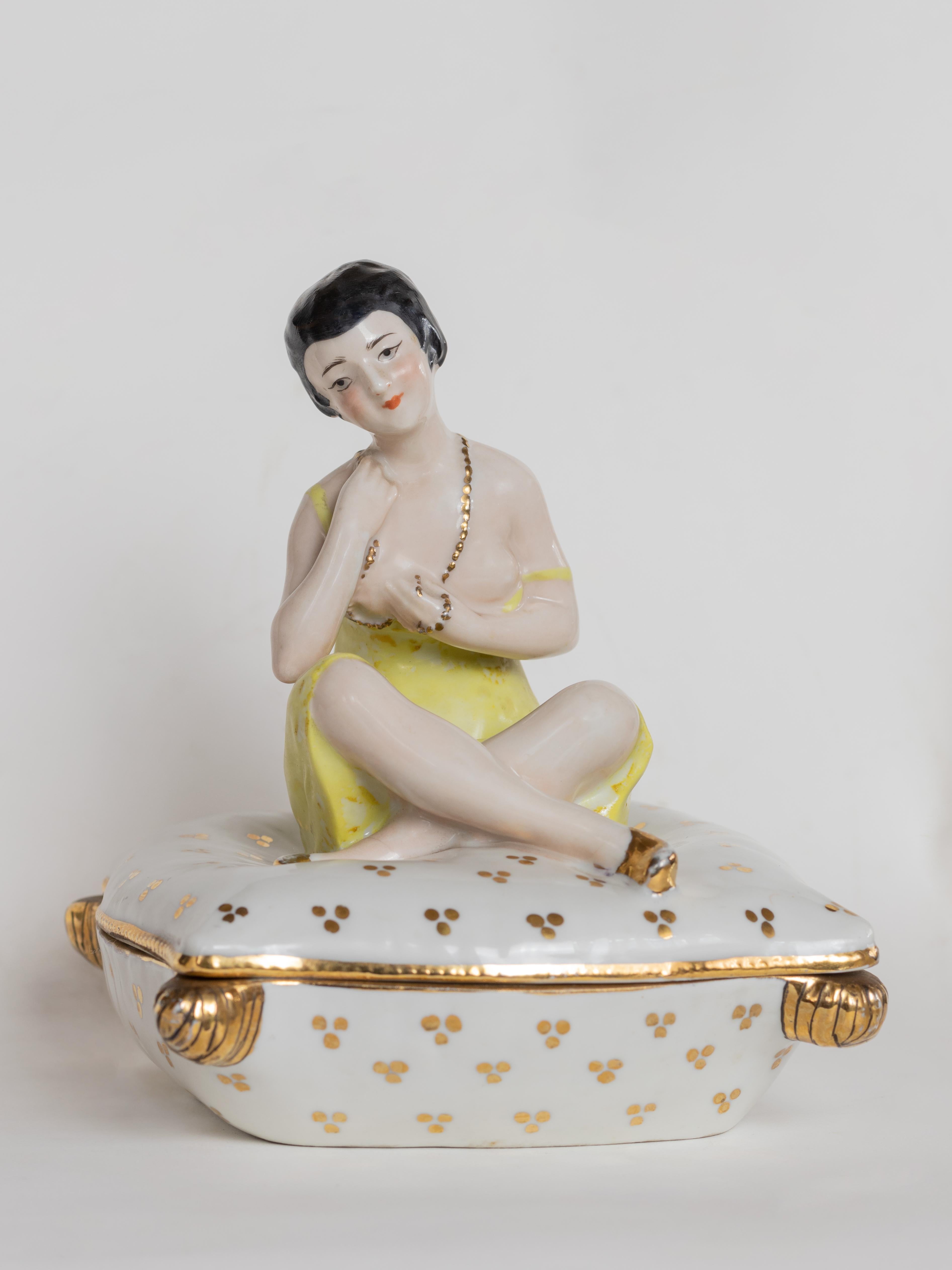 Diese Art-Déco-Porzellandose zeigt die Figur einer sitzenden Frau in einem leuchtend gelben Kleid, die anmutig auf einem weißen Kissen sitzt, das mit zarten Goldmotiven und Eckmuschelfüßen verziert ist. 

Als Puderdose oder Schmuckkästchen
