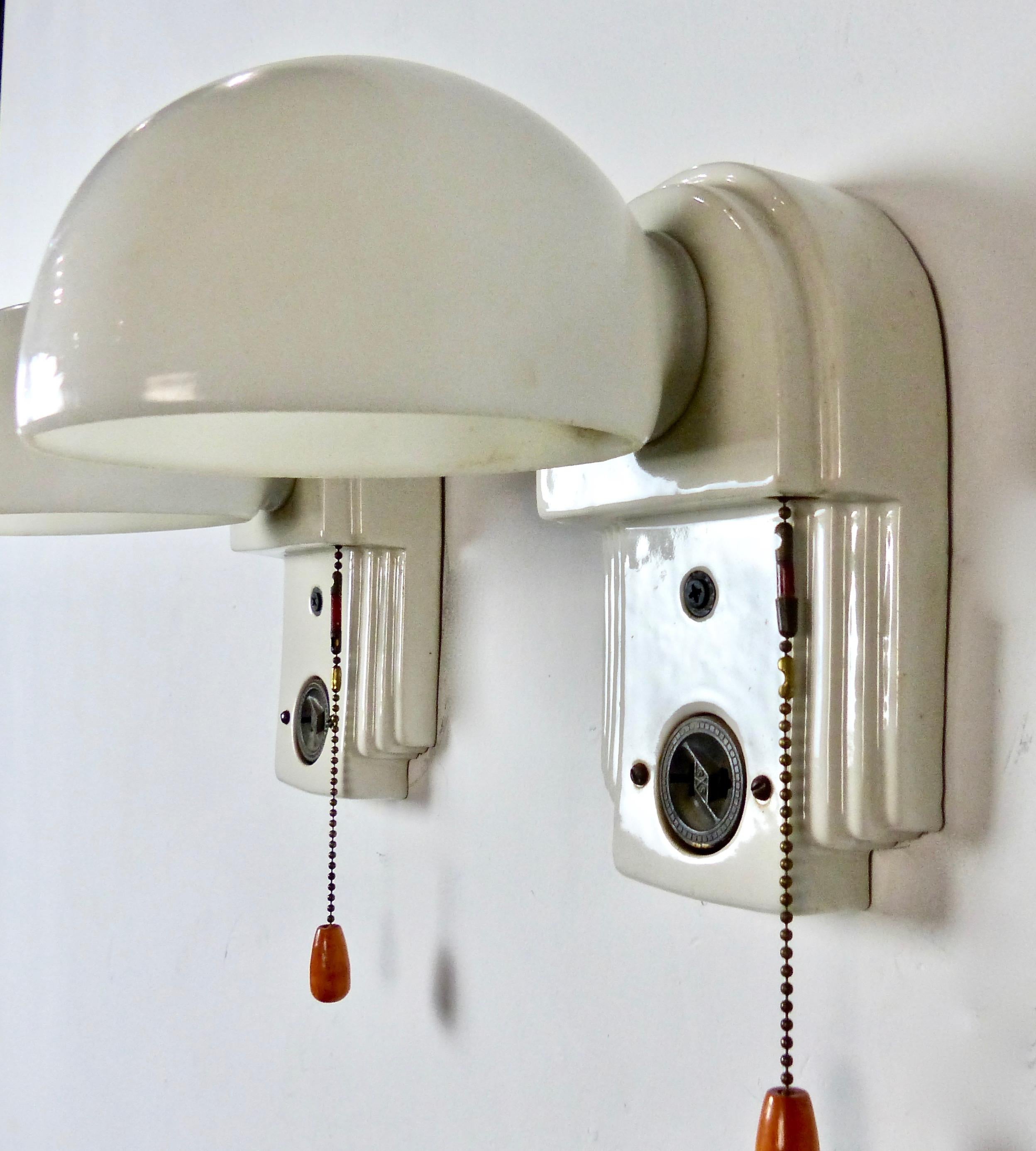 A matched pair of charming white porcelain wall sconces with milk glass shades from circa 1920. Pull-chain on/off switch with wooden knob. Convenient extra electrical outlet incorporated into the design. Re-wired and CSA approved to current