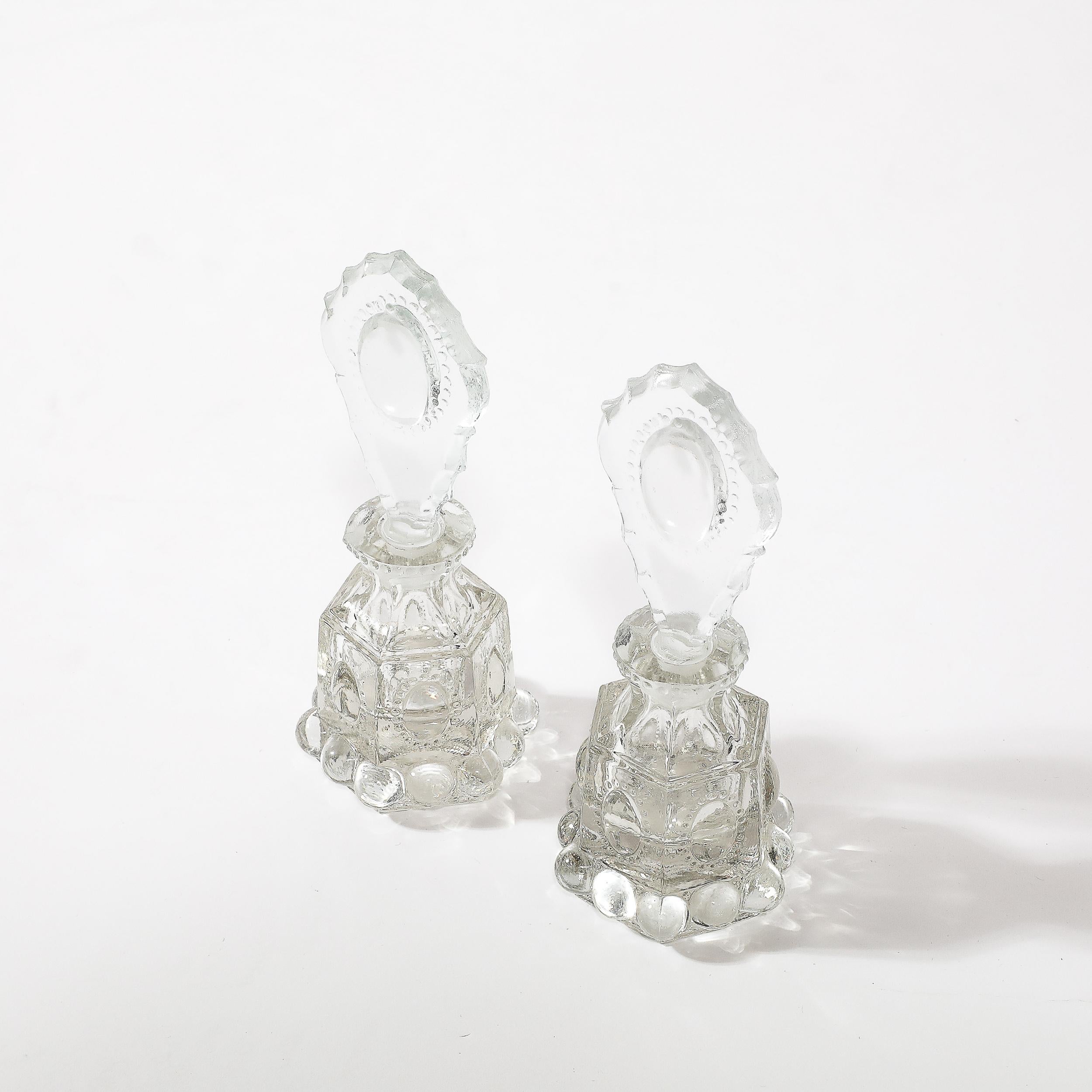 Cette exquise paire de porte-parfums en verre pressé de style Art déco illustre à merveille l'élégance et le savoir-faire du design américain des années 1930. Chaque porte-bouteille est orné de détails complexes le long du corps, ajoutant texture et