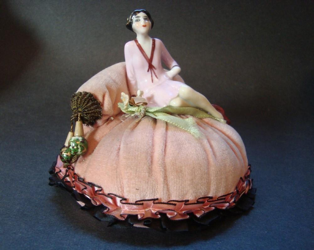What a beauty this pincushion is, the full figured flapper sitting casually on a peach coloured velveteen pin cushion. It has a pillow for her to lean on and that has corded tassels at each end. There is two-toned silk coloured ribbon around the