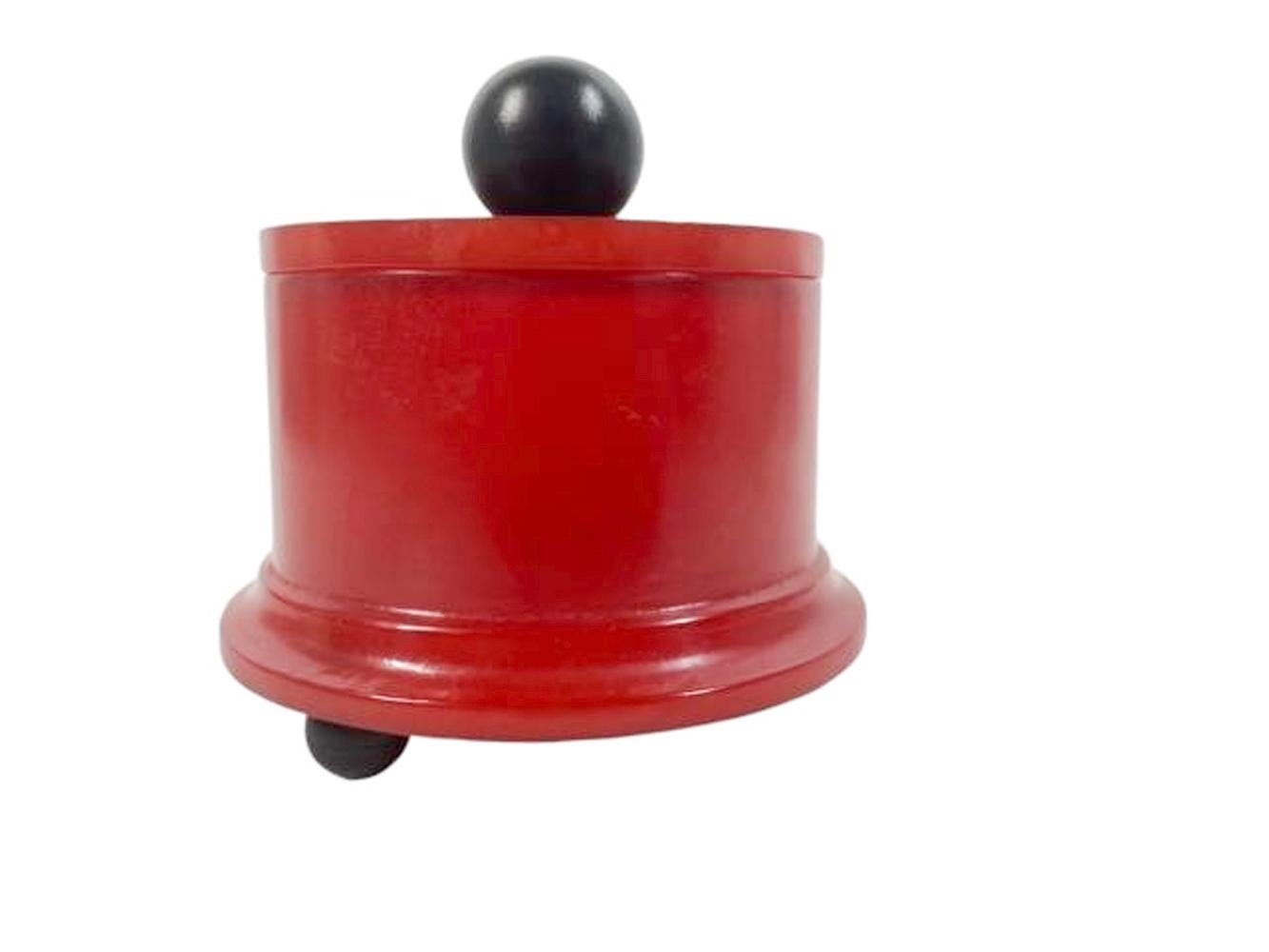 Caja de escritorio o tocador Art Decó de baquelita rojo cereza, de forma cilíndrica, con base escalonada moldeada y tapa conforme con pomo de bola de baquelita negra, elevada sobre tres patas de bola de baquelita negra.