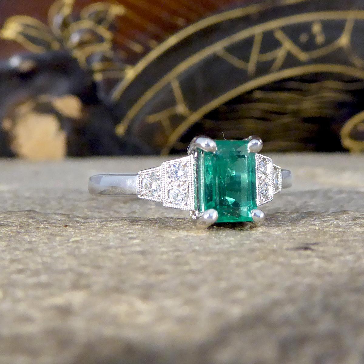 A beautiful ring has been created and crafted in an Art Deco style but is new and never worn. It has been designed and carefully crafted with a vibrant and bright Emerald in the centre in a four claw setting with two milgrain set Diamonds tapering