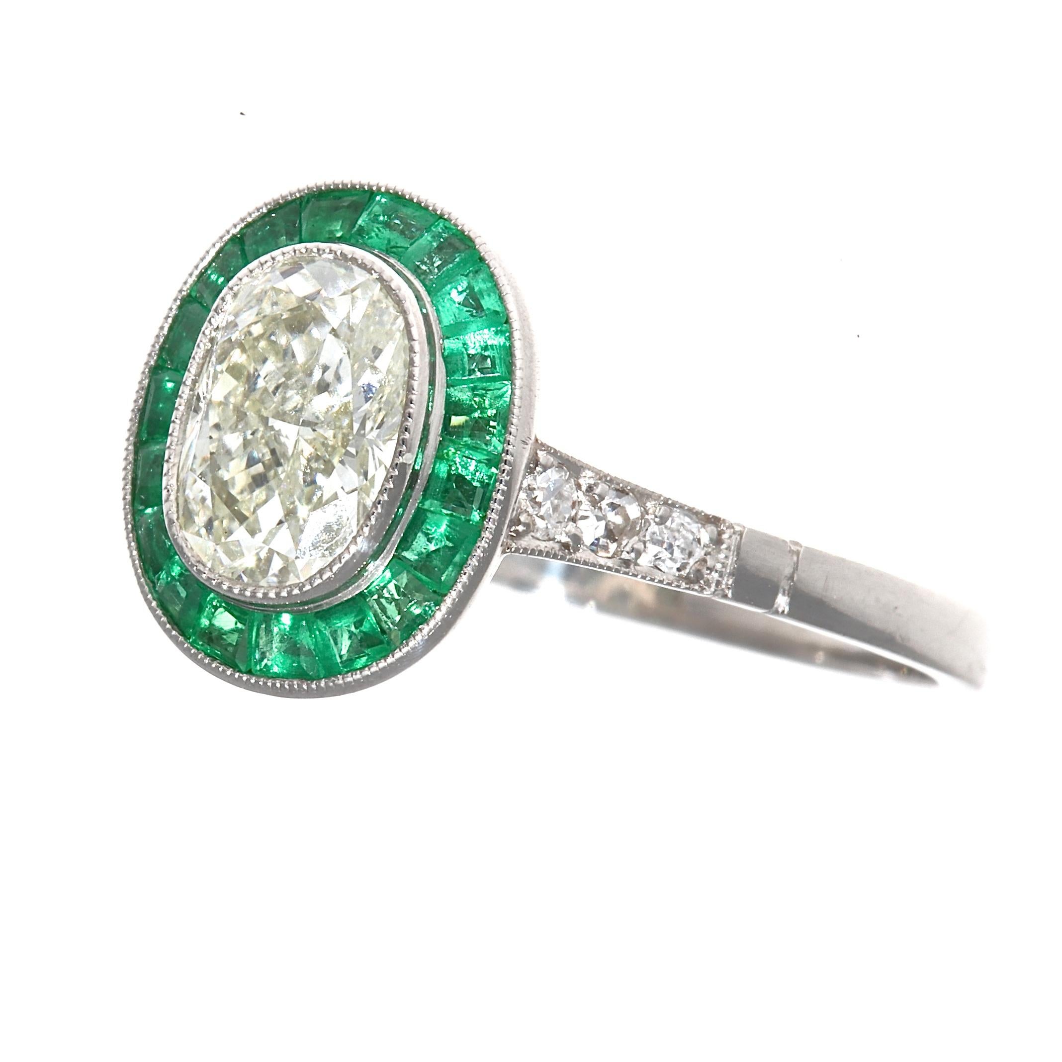 A design driven by colorful passion giving devotion to the art deco time period. Featuring a 1.01 carat oval cut diamond that is L color, SI1 clarity. Brilliantly surrounded by a vibrant green halo of calibrated emeralds specially cut to mirror the