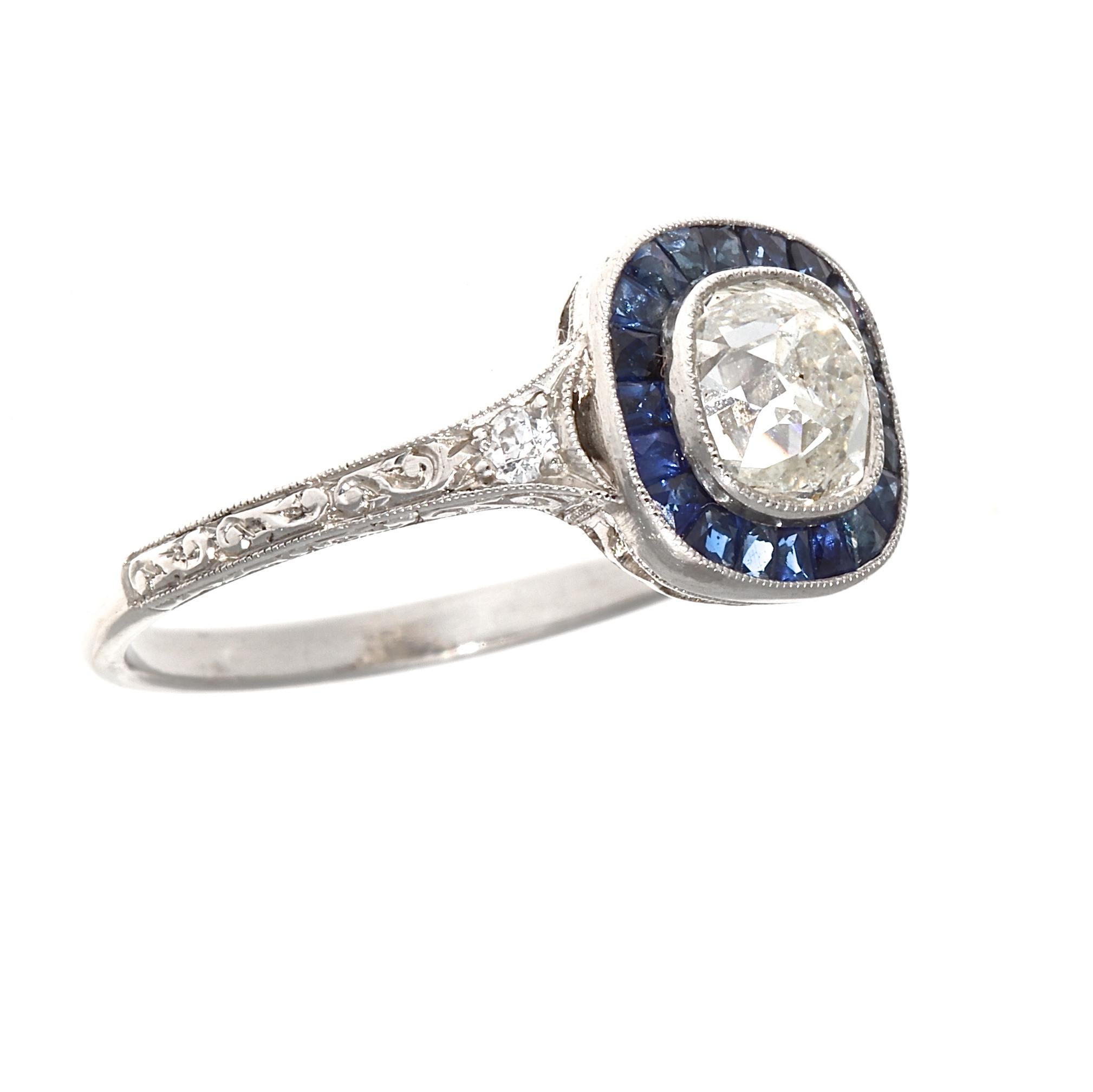 Steeped in the tradition of the romantic and elegant art deco era. Featuring a 1.07 carat old mine cut diamond that is I color, SI2 clarity. Surrounded by a radiating halo of royal blue sapphires. Hand crafted in platinum. Ring size 7 and can easily