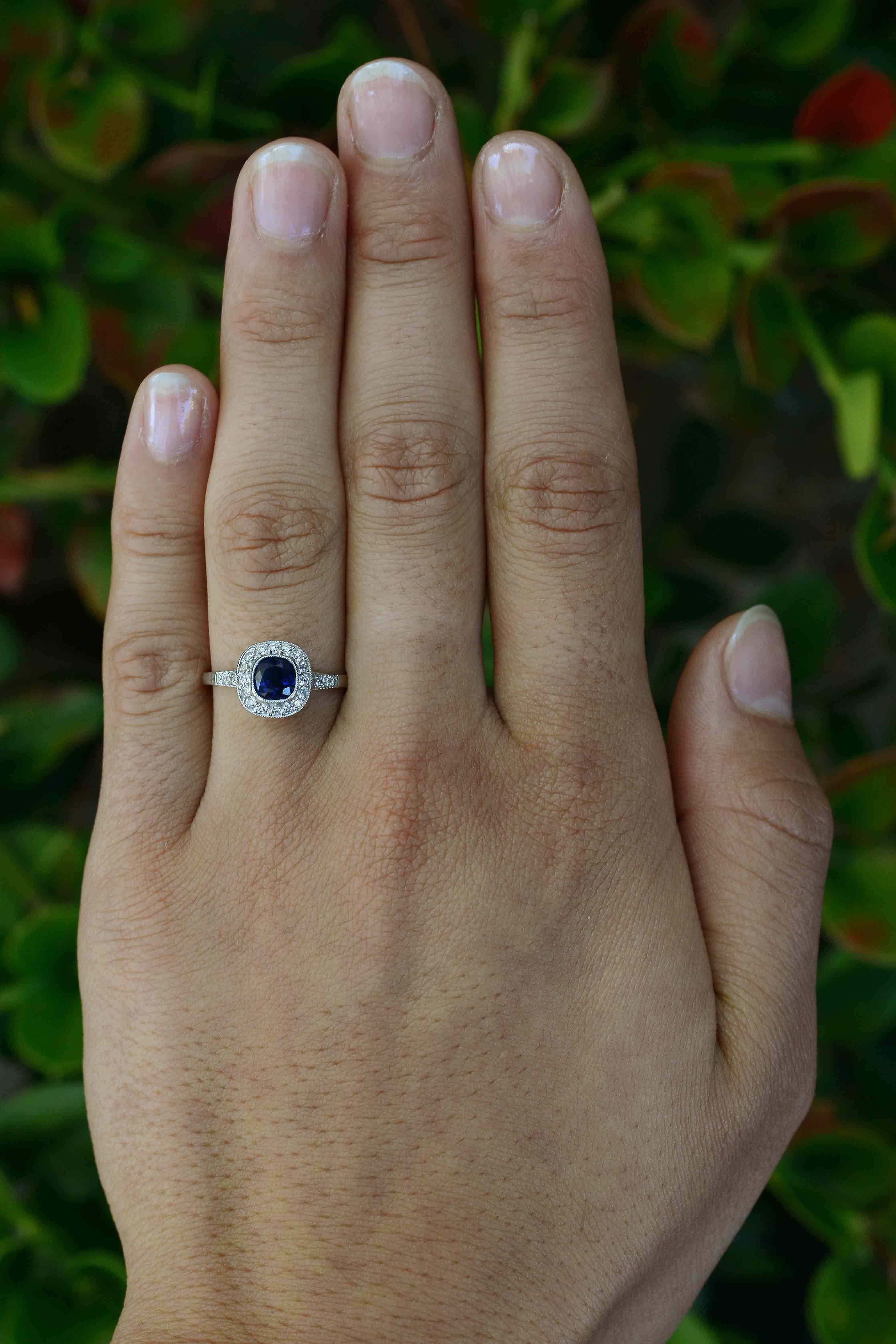 Crafted of everlasting platinum and set in a milgrain bezel is a rich and luminous antique blue sapphire that floats within a shimmering diamond halo. A wonderful and desirable cushion cut gemstone engagement ring. Supporting the filigree, scrolled