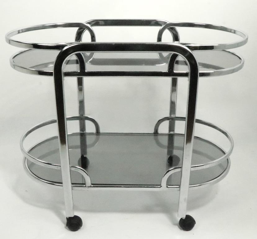 Oval serving, bar cart in the Art Deco Revival style, circa 1970s-1980s. The cart is oval, it features a removable upper level tray, and wheel castor feet. The top tray is removable, perfect for in home entertaining
Original tinted glass shelves,