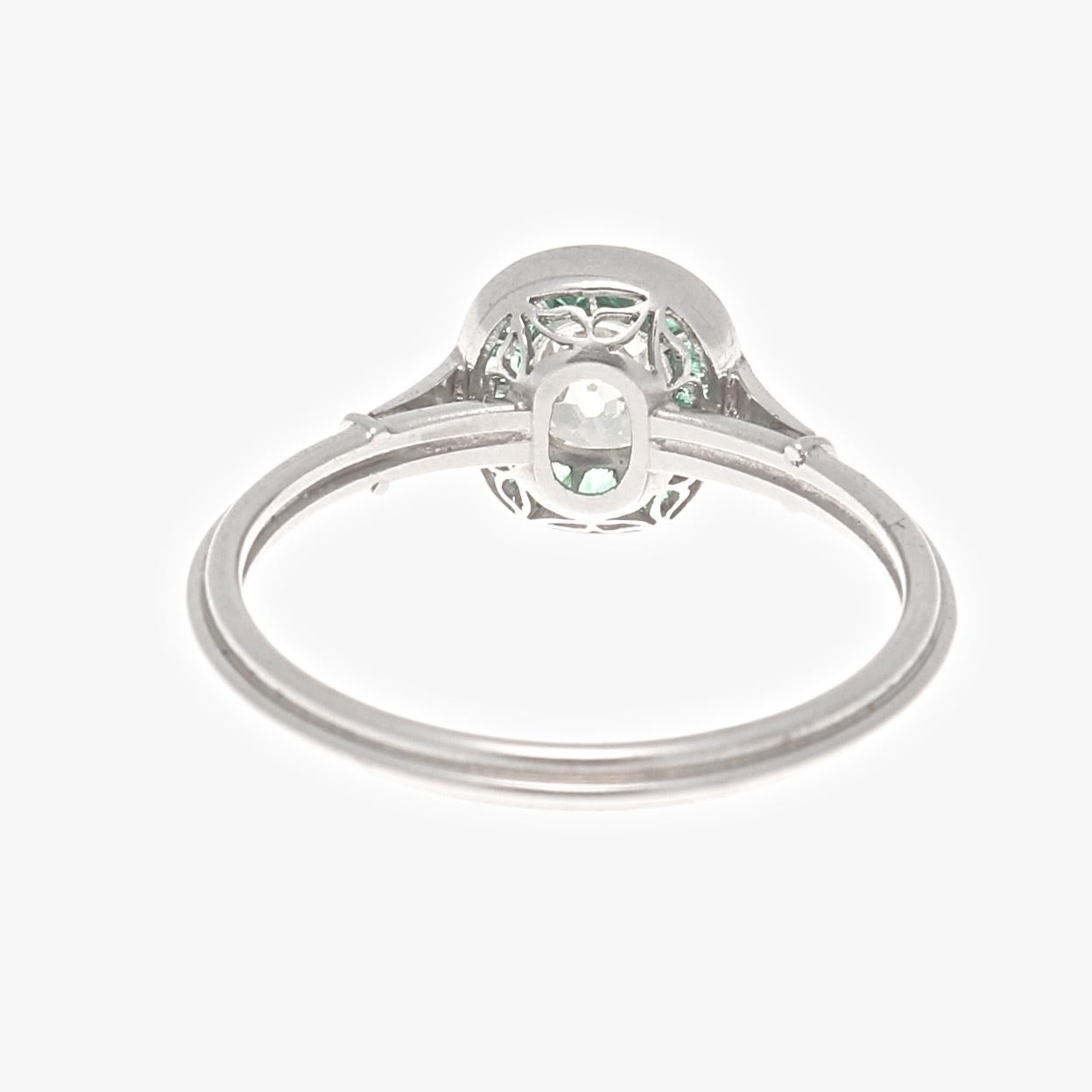 A design driven by colorful passion giving devotion to the art deco time period. Featuring a 0.66 carat old mine cut diamond that is L color, VS1 clarity. Brilliantly surrounded by a vibrant green halo of calibrated emeralds specially cut to mirror