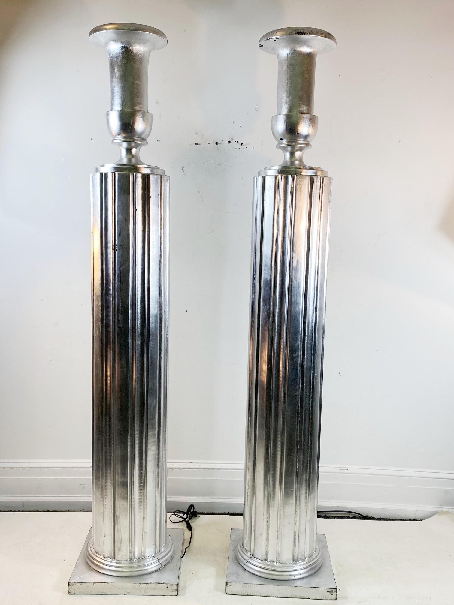 Art Deco Style pair of monumental torchère floor lamps consisting of fluted column bases with a large urn mounted on top. The pair is made of carved and silvered wood. In great vintage condition with age-appropriate wear and use.