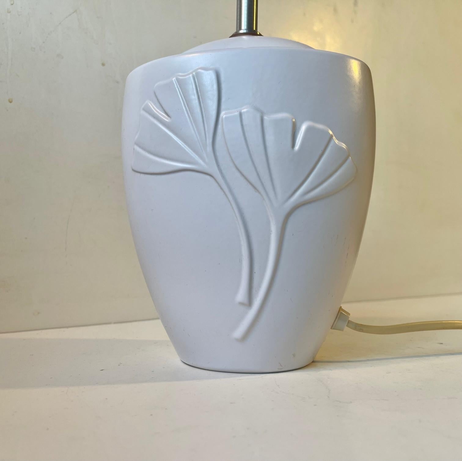 A white glazed table light featuring stylized relief flowers and fluted white shade. Art Deco revival in style. Made at Søholm in Denmark circa 1970-80. Measurements: H: 40 cm, Diameter: 30 cm. 
For the US. It will come installed with a 110 watt