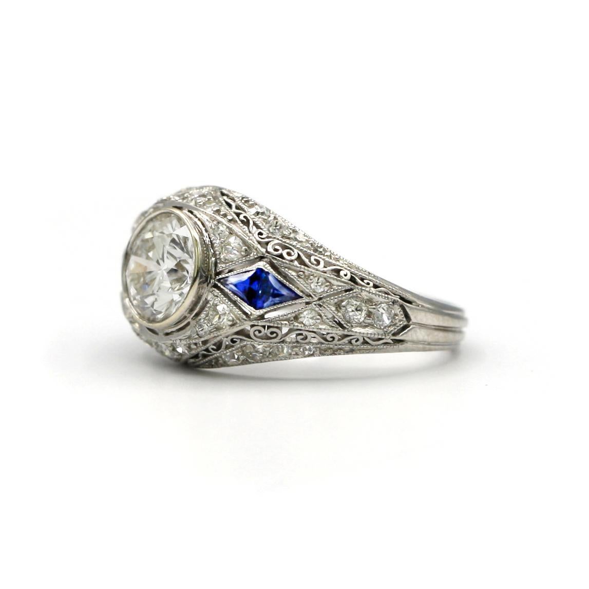 This beautifully detailed 18 Karat White Gold Art Deco ring features a center white round diamond, weighing approximately 1.20 carats, 38 round white diamonds weighing approximately 0.40ct and 4 triangle shaped sapphires weighing approximately
