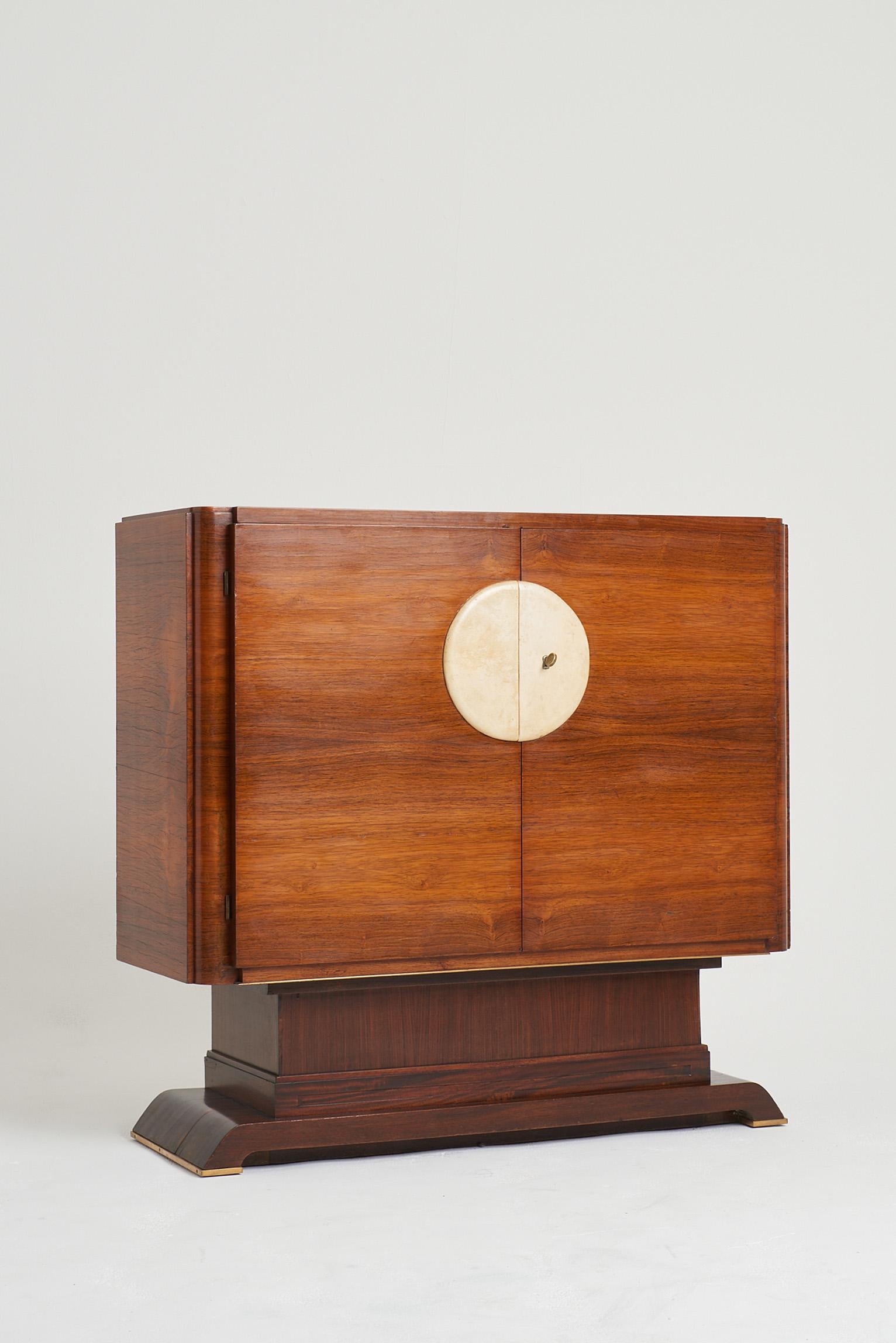 An Art Deco walnut veneered cabinet, of unsual shape, the two doors adorned with a large velum disk and brass key, opening to reveal a central adjustable shelf and two mahogany veneered drawers.
France, Circa 1930.