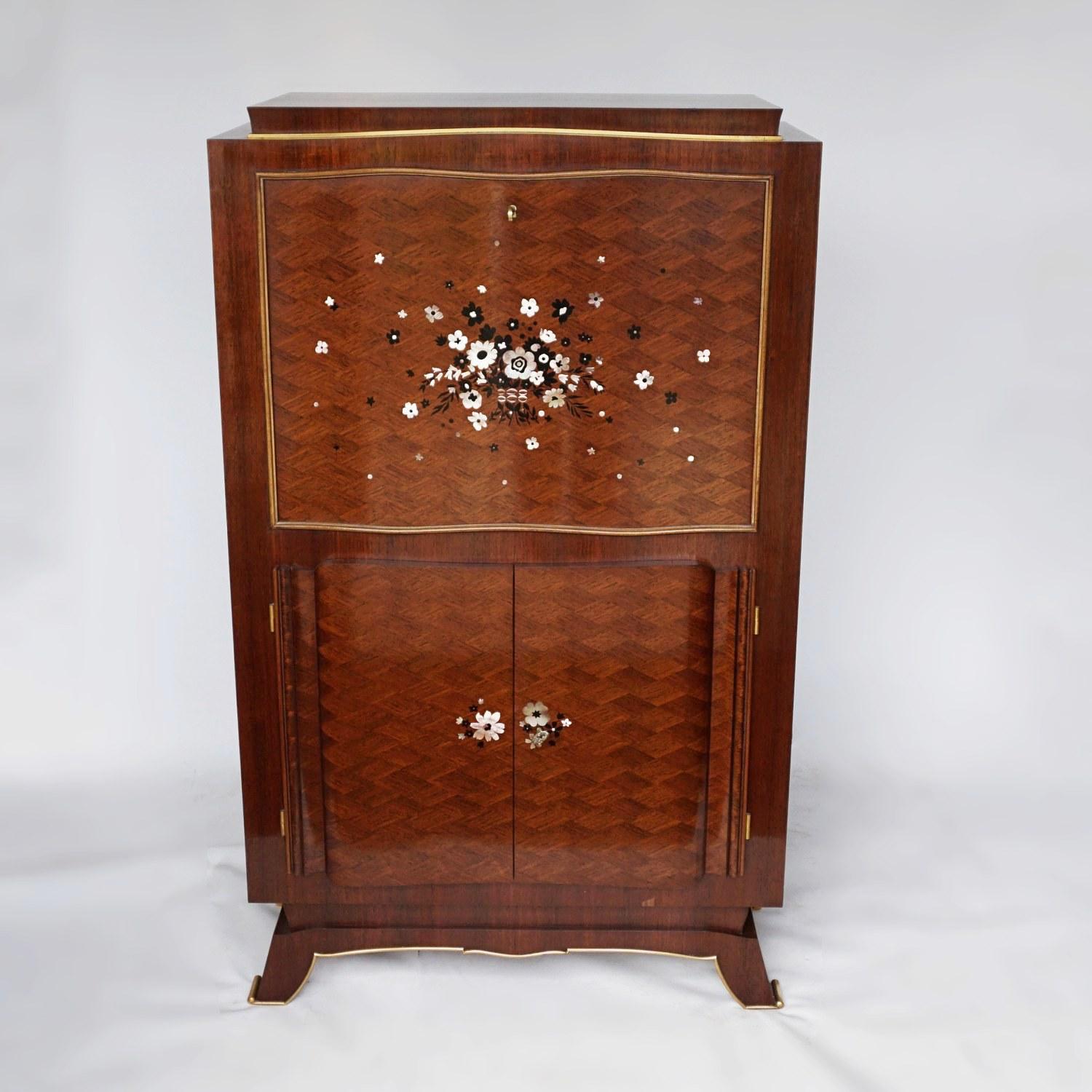 An Art Deco rosewood marquetry cocktail cabinet by Jules Leleu, (1883-1961). Stepped top over a parquetry and marquetry inlaid drop-front cocktail cabinet, with ebony and mother of pearl inlay flower decoration. Mirrored lit interior to top, with a