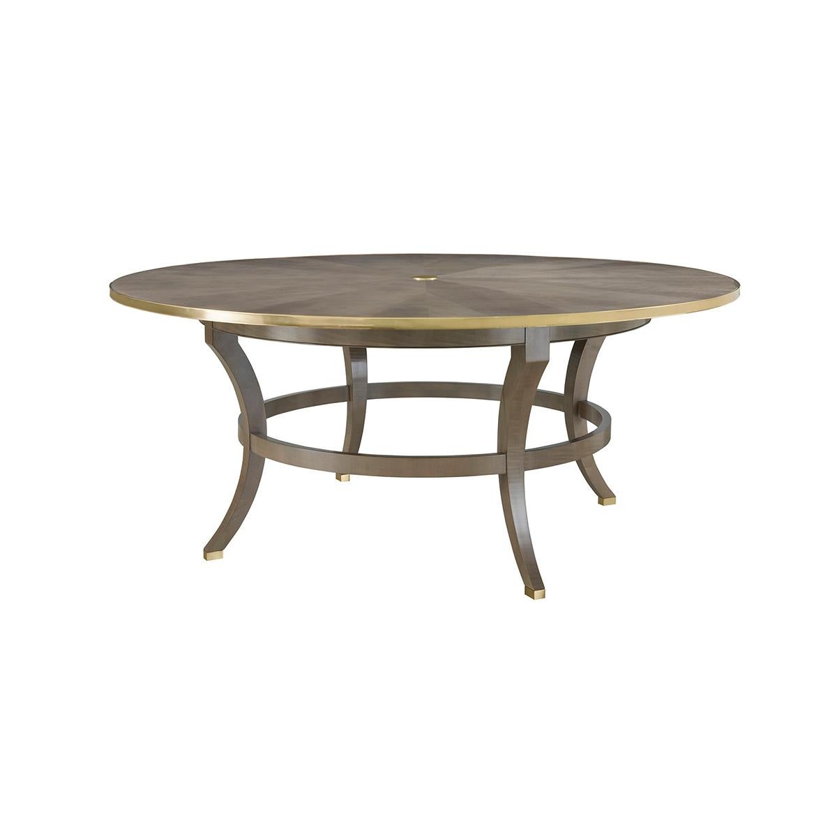 An Art Deco round dining table. Greyed Sycamore veneers with a rayed inlaid top, a solid brass central applique and brass trim molding. Raised on four sabre form legs with a round stretcher and brass feet.

Dimensions: 72