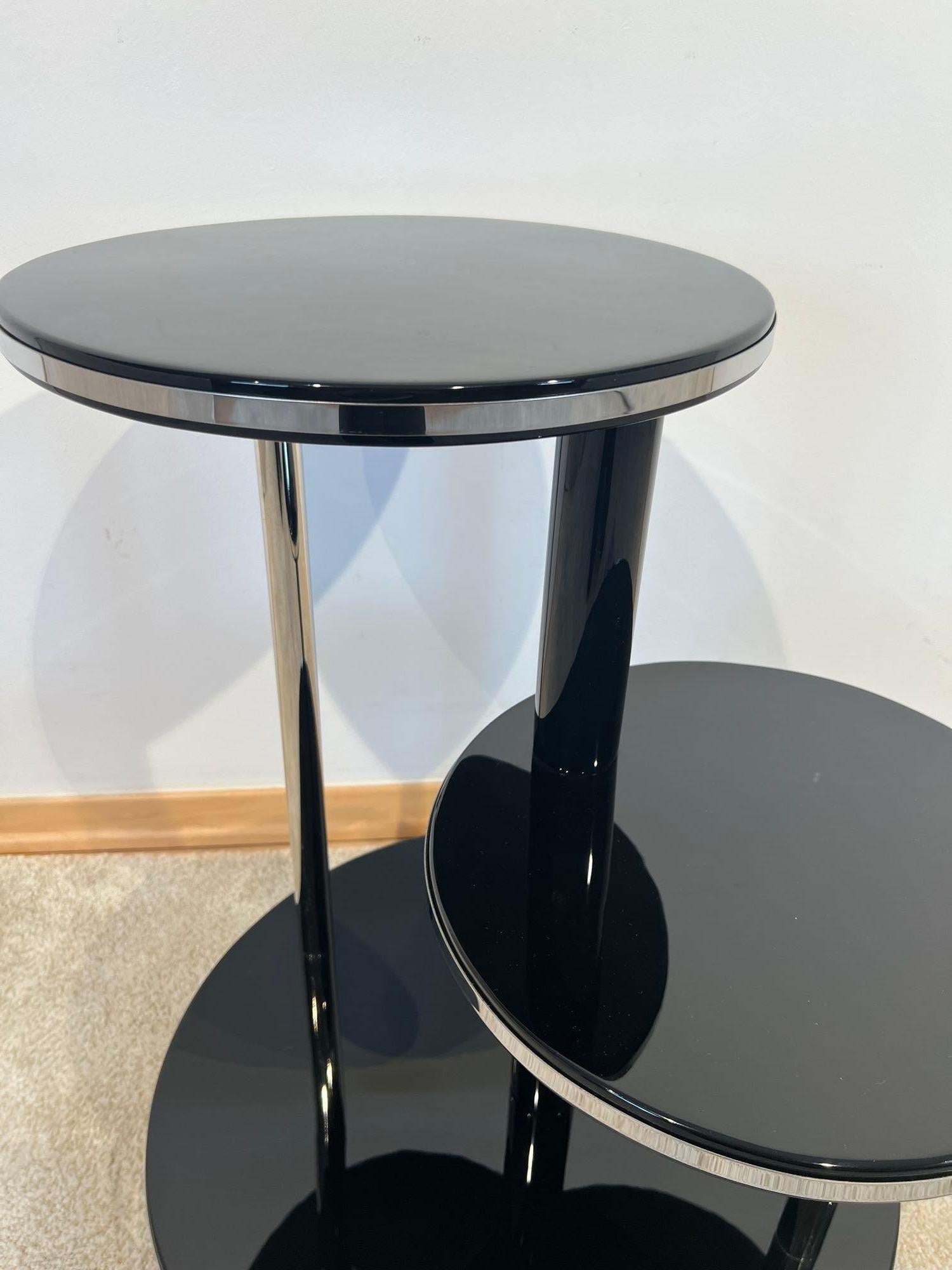 Round Art Deco side table, flower stand or three-tier etagere table
Black piano lacquer on wood, lacquered and polished to a high gloss. Two vertical chrome-plated metal rods and a black painted stele in the middle. All three levels have a stainless