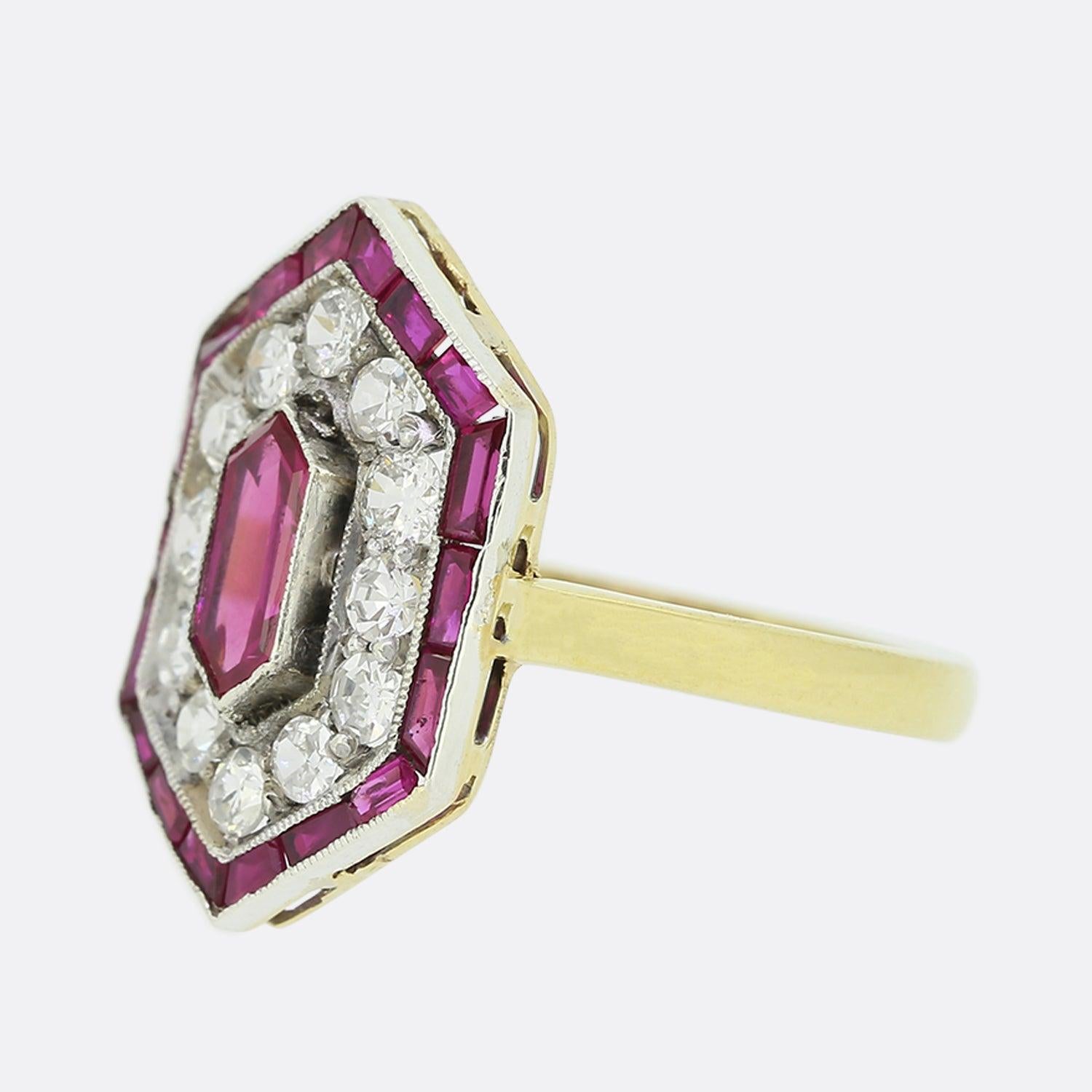 This is an Art Deco 18ct yellow gold ruby and diamond cluster ring. The ring has been crafted in a hexagonal shaped setting and is set with a central ruby which is surrounded by a halo of old and single cut diamonds and then a border of calibre cut