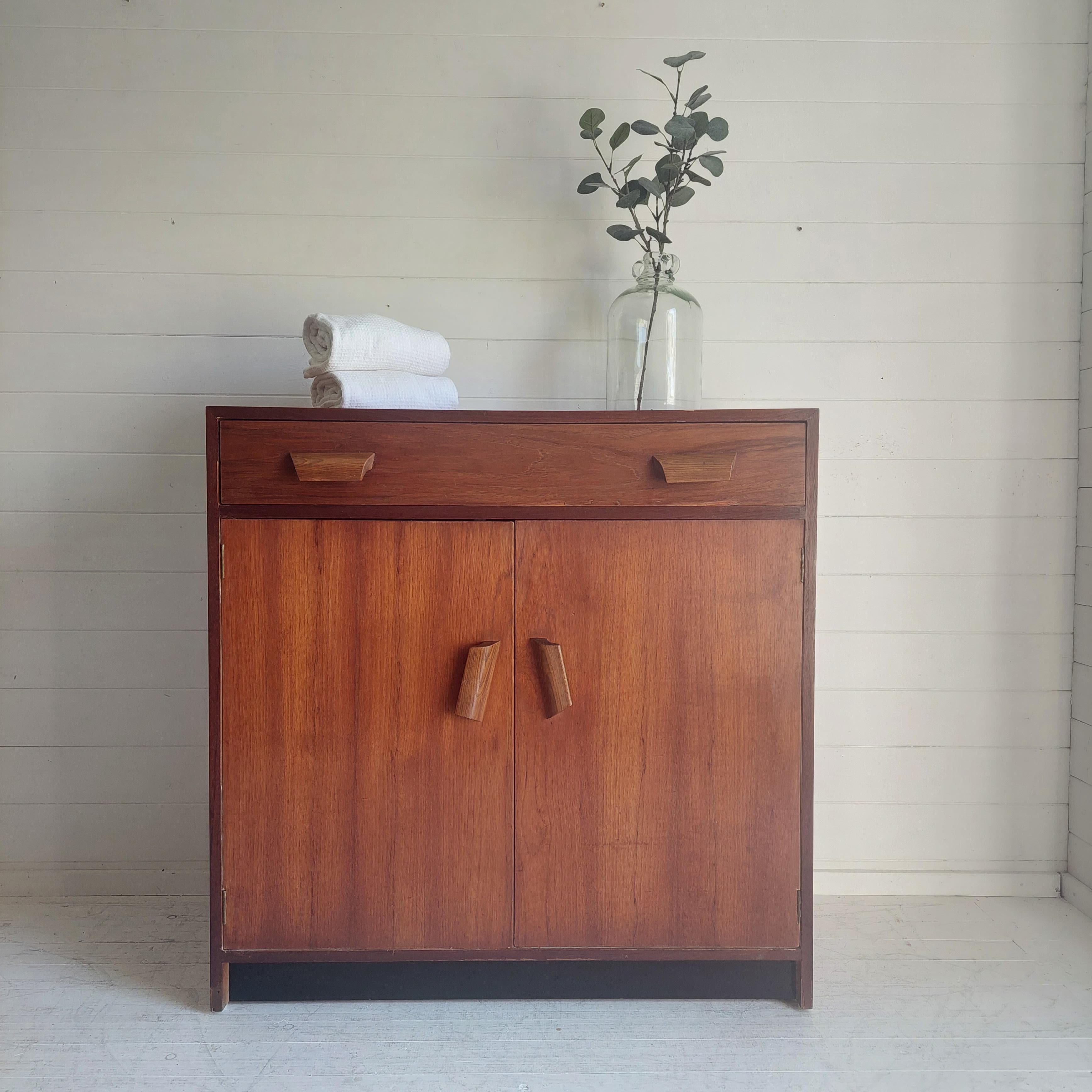 A Late Art Deco early Mid Century cupboard 50s

A beautifully simplistic, rustic look teak shelved storage cupboard with drawer.
In contrasting rich teak with  huge carved oak handles with a two door cupboard hosting a shelf above a single drawer