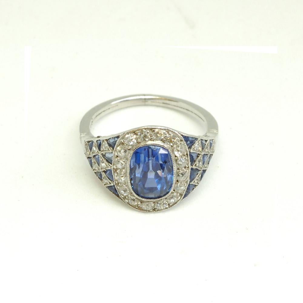 An Art Deco sapphire and diamond cluster ring of oval shape. The triangular shaped shoulders are set with alternating sapphires and diamonds in a stylised checker-board pattern. Mounted in millegrain edged platinum throughout. English, circa
