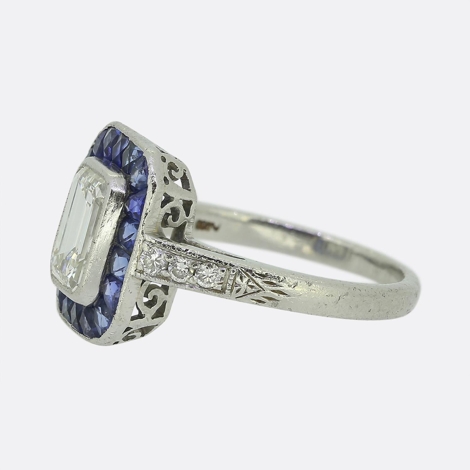 Here we have an outstanding platinum cluster ring crafted at a time when the Art Deco style was continuing to revolutionise the world of design. A remarkably clean, bright white emerald cut diamond sits slightly risen at the centre of the face. This