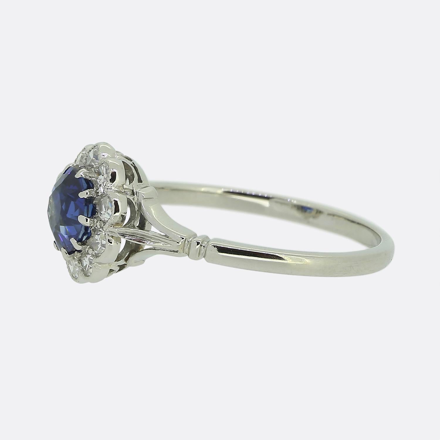Here we have a sweet and charming cluster ring dating back to the Art Deco movement. This elegant piece has been crafted from 18ct white gold and features a single round faceted sapphire at the centre of the face. This principal stone possesses a