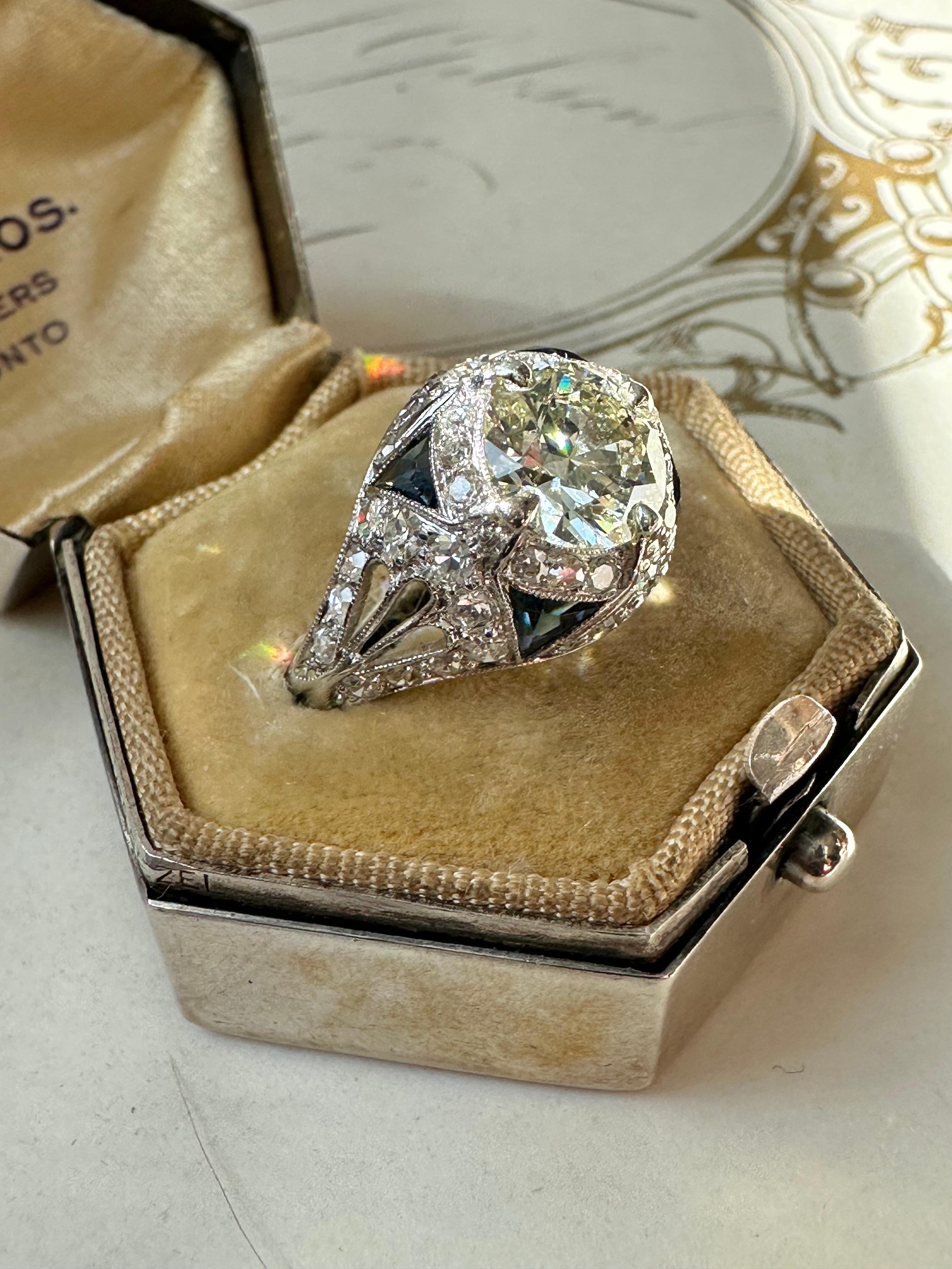 This spectacular Art Deco ring boasts a lively 1.53 carat transitional-cut diamond wrapped in a halo of glittering single-cut diamonds and crowned by four deep blue synthetic calibre sapphires. Expertly hand fabricated in platinum, the shoulders are