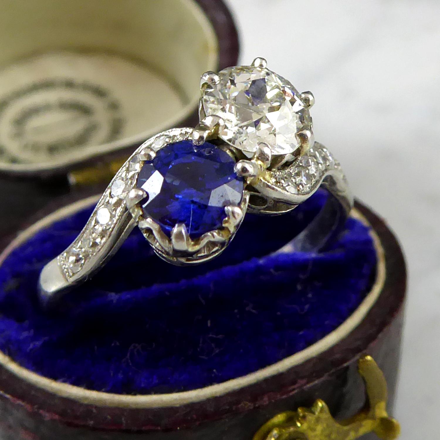 An Art Deco vintage ring in a two stone cross over twist style set with an old European cut diamond and a round, mixed cut sapphire each in a six claw basket setting.  The diamond measuring approx. 5.42mm x 5.50mm x 3.35mm deep, calculated to weigh