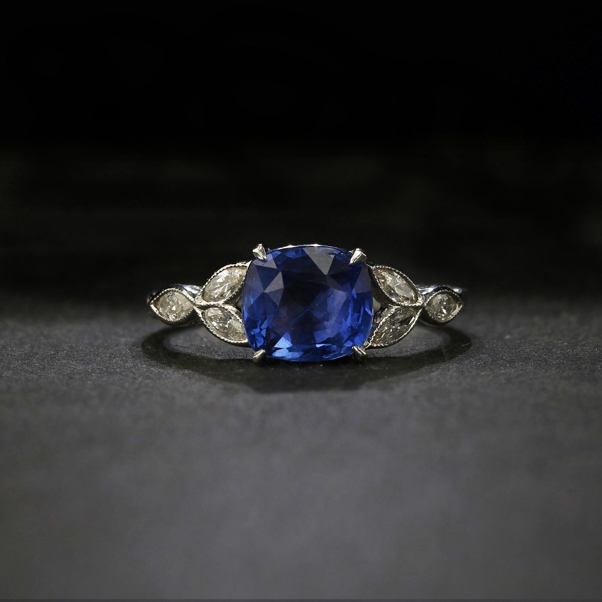 The rich blue of this cushion-shaped sapphire, weighing in at over two and a half carats, is delicately framed by a half dozen twinkling white marquise diamonds in fine millegrained bezel settings, all set against gleaming 14k white gold in an