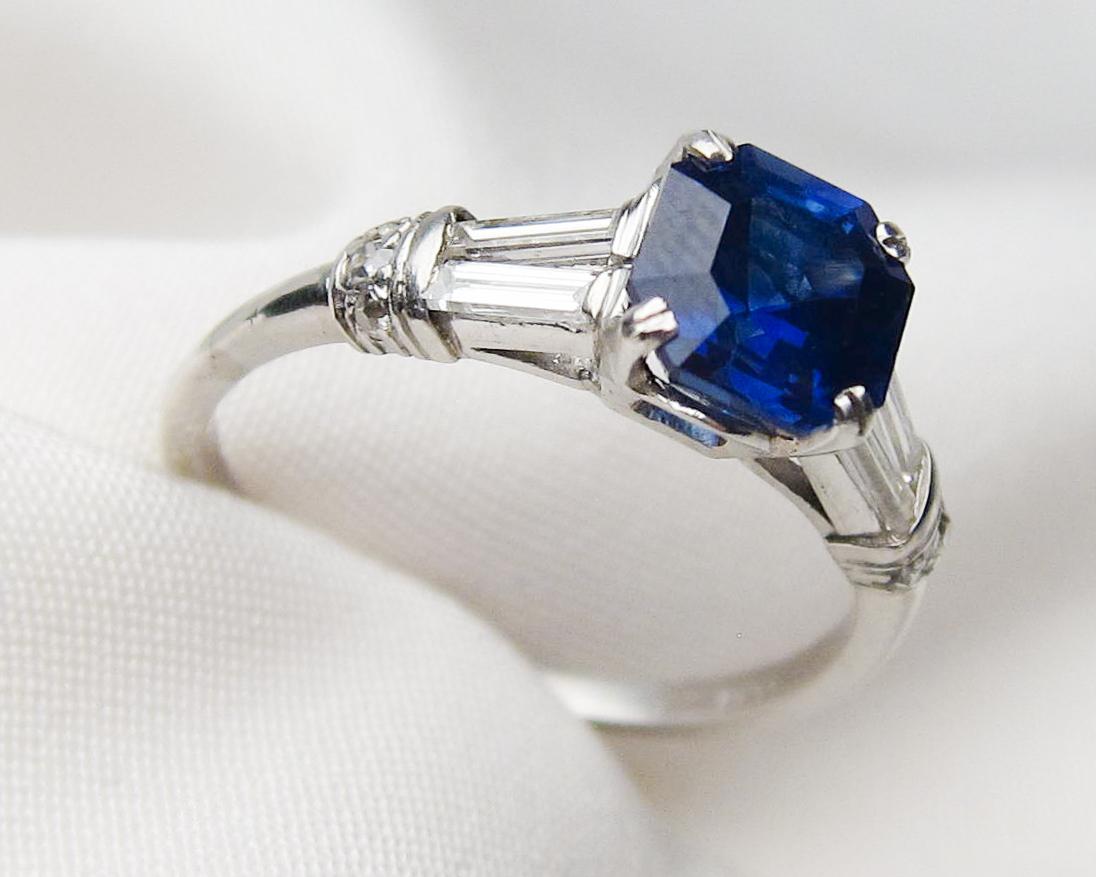 Circa 1930. This stunning platinum Art Deco Era ring features a central square emerald-cut natural blue sapphire weighing 2.25 carats. The stone is lightly included with a violet-blue hue and dark saturation. Accenting each side of the central stone