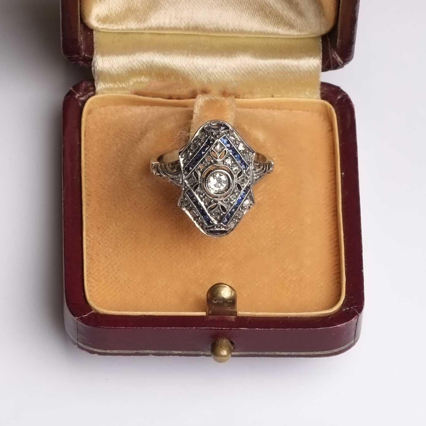 Art-Deco-Ring aus 18-karätigem Gold mit Diamanten im Rosen- und Altschliff sowie Saphiren im kalibrierten Schliff.

Höhepunkte
- Art Deco
- 18ct Weißgold
- Diamanten im Alt- und Rosenschliff
- Saphire

Dimensionen
- Länge 2,8 cm x Breite 1,2 cm
-
