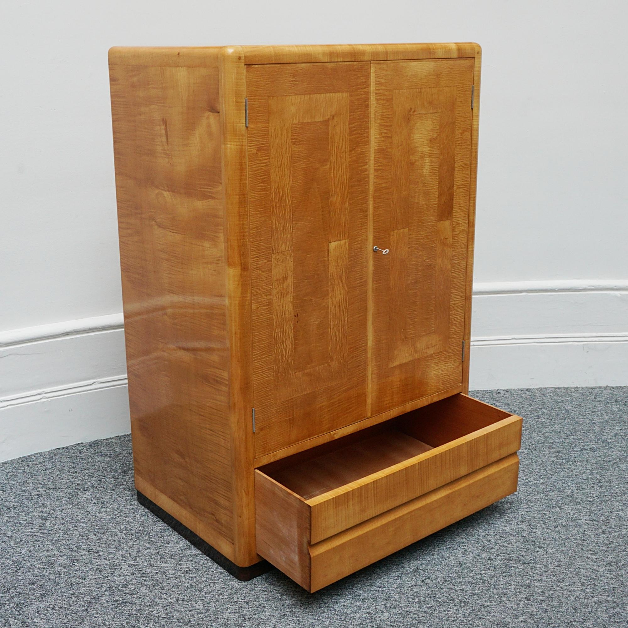 An Art Deco cabinet by Betty Joel. Upper cabinet section opens to reveal a hanging rail with six shelves to the side and a tie rack to inside of door. Lower single drawer. Satin birch veneered with Macassar ebony banding to base. Designer and makers