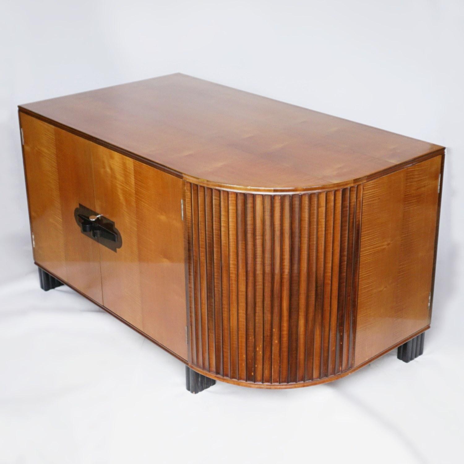 An Art Deco desk by Maurice Adams. Satinwood veneered on solid mahogany with ebonised fluted feet and handles. Front section of five drawers with a shelved cupboard. Large shelved cupboard section to back with a curved tambour feature.