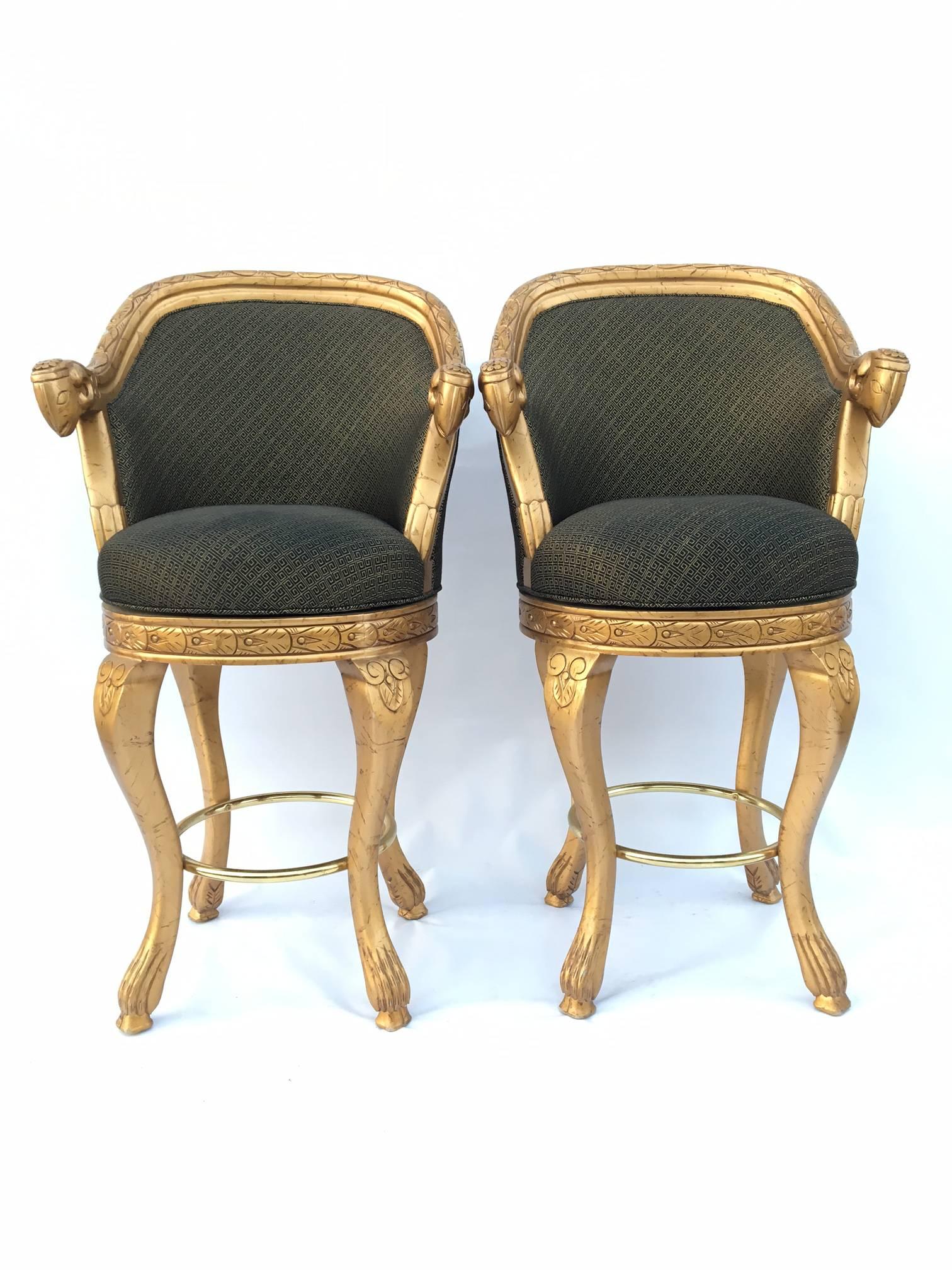 Pair of gold neoclassical Savoy style bar stools featuring a Greek key print upholstery and carved ram's head arms. Swivel seats and brass foot rails. Excellent vintage condition with only very minor signs of use and slight darkening of upholstery.