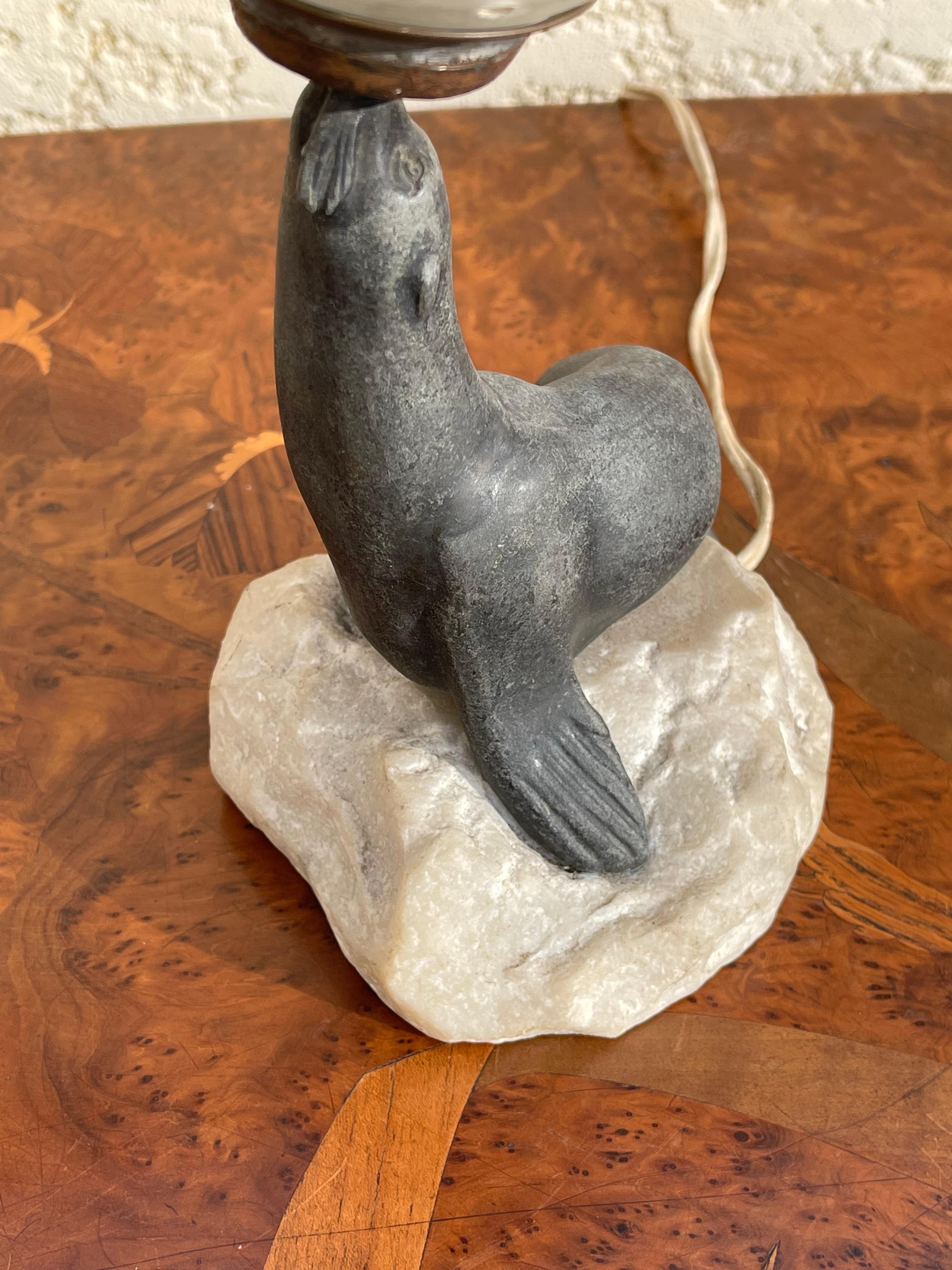 Art Deco lamp depicting a samac sea lion sitting on a white marble rock holding a glass globe on the tip of its snout. It is in good condition.

Dimensions
Rock 12cm x 11cm
Total height 26 cm