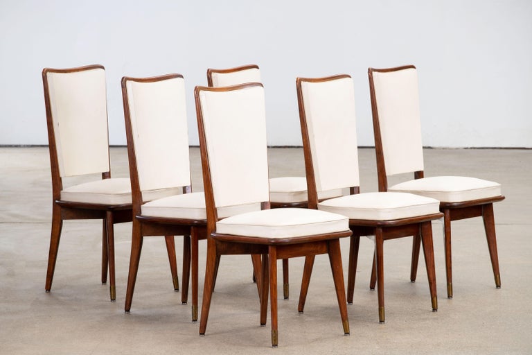 Set of six upholstered high back chairs covered in white vynil, exhibiting traditional French design elements in a deep oak finish.

In the style of René Prou, Albert-Lucien Guenot, Pomone, André Arbus, Baptistin Spade, Charles Dudouyt, Herman