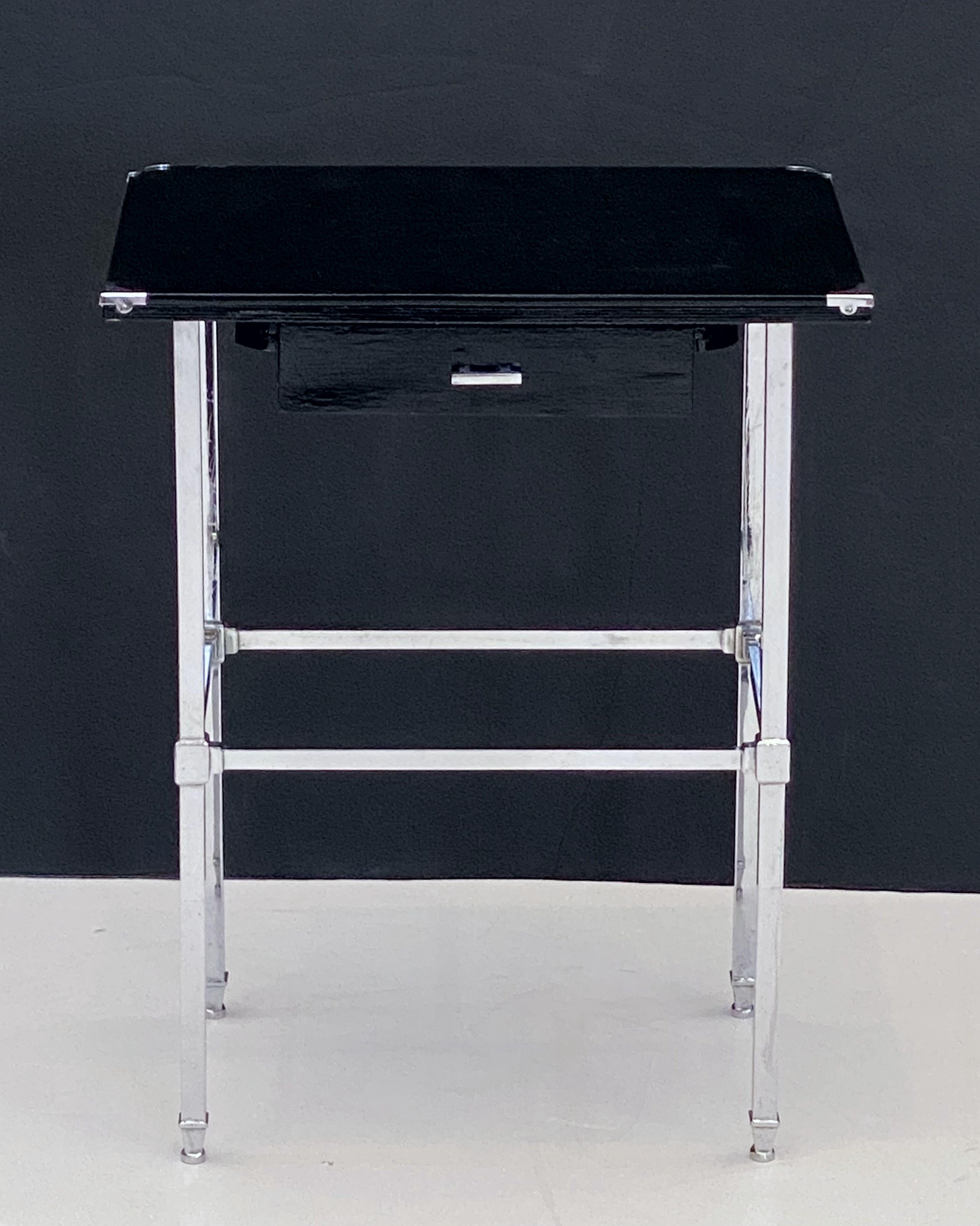 A fine English side or end table of black-lacquered wood and chrome, in the Art Deco style, featuring a chrome-bound rectangular top, over a frieze with drawer and chrome handle, resting on a four-legged support of chrome.