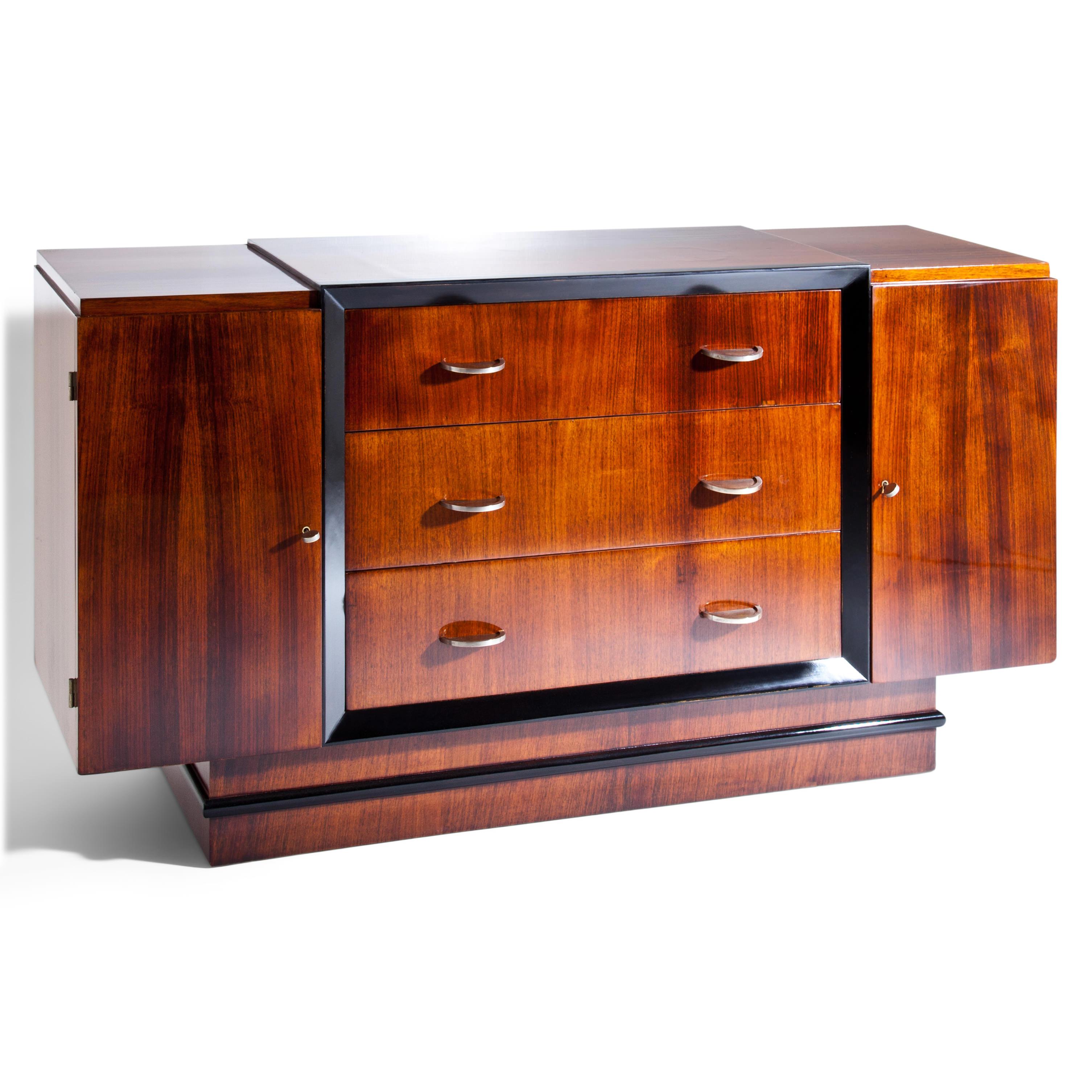 Art Deco sideboard with three drawers and two doors on a straight plinth with ebonized all-round profile strip. The disc-shaped brass handles emerge as semicircles from the veneered fronts. The drawers are visually set off by an ebonized frame.