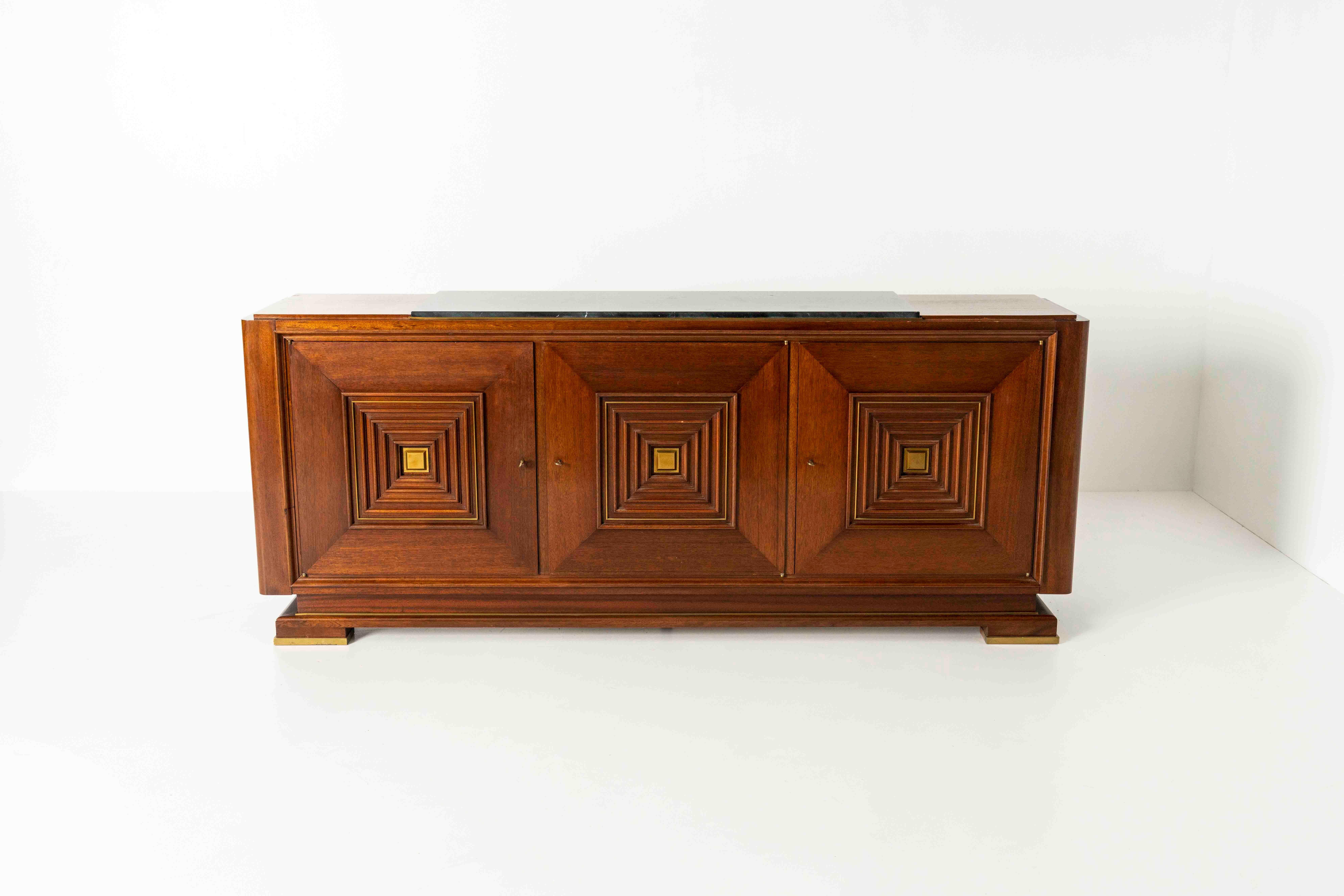 Sideboard in the Style of Maxime Old in Mahogany, Brass, and with a Marble Top. It has a drawer in the middle and shelves behind the left and right doors. We especially love the design of the legs and feet with the brass details. This impressive