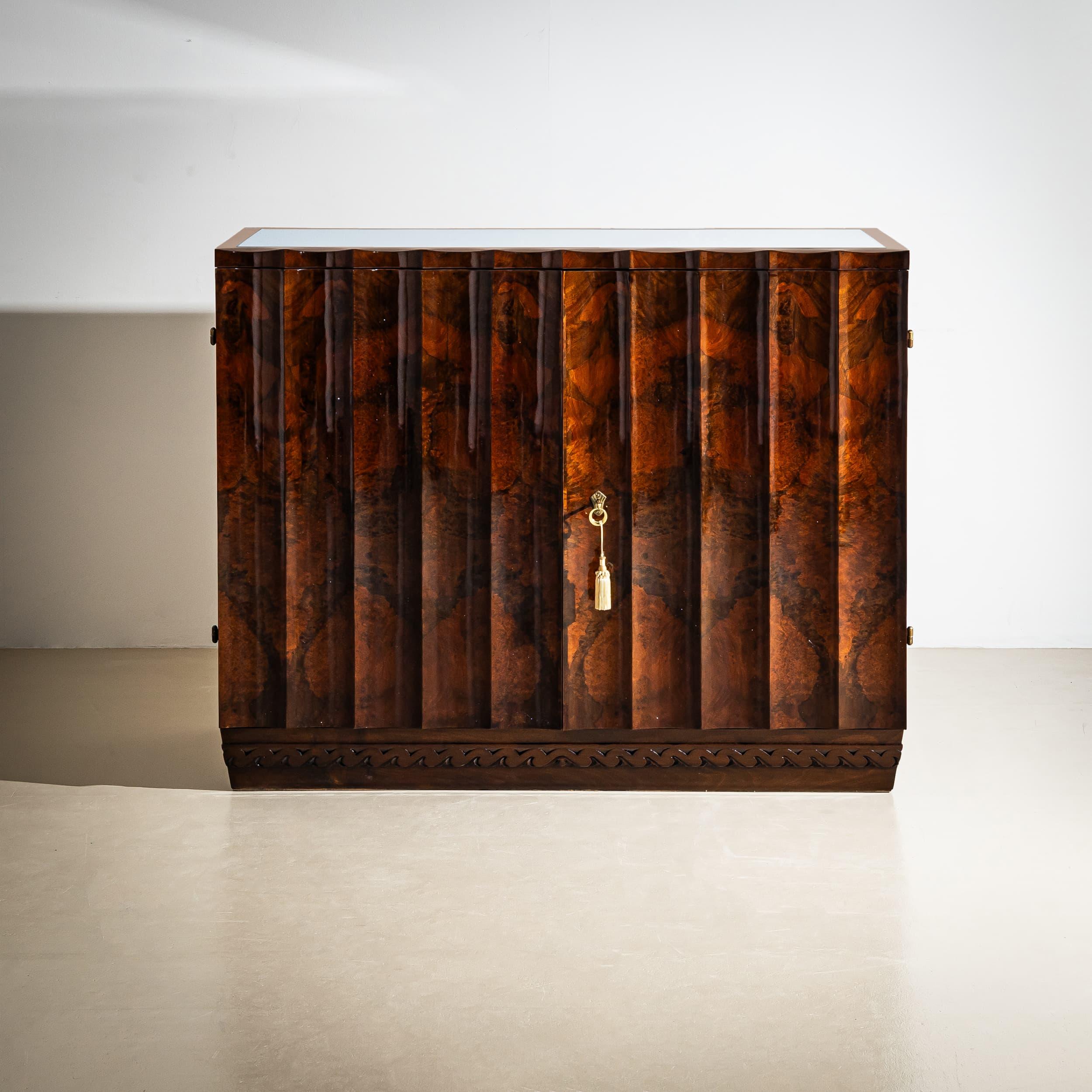 Aparador de dos puertas en nogal con un distintivo frente curvo y un friso ondulado a juego a lo largo de la base de zócalo recto. El diseño dinámico se refleja en el meticuloso patrón de chapa de madera de grano vivo. El interior está equipado con