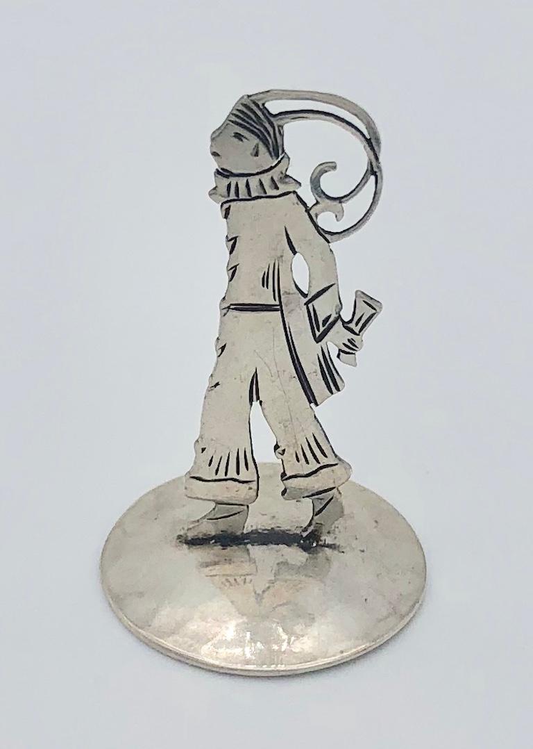 Kleine Figur einer modischen Dame aus den 1920er Jahren, die einen Hosenanzug, einen Hut mit Federn und Handschuhe trägt. Er steht auf einem Sockel aus gehämmertem Silber. Die Figur ist auf beiden Seiten eingraviert. 

Sie ist mit den Initialen